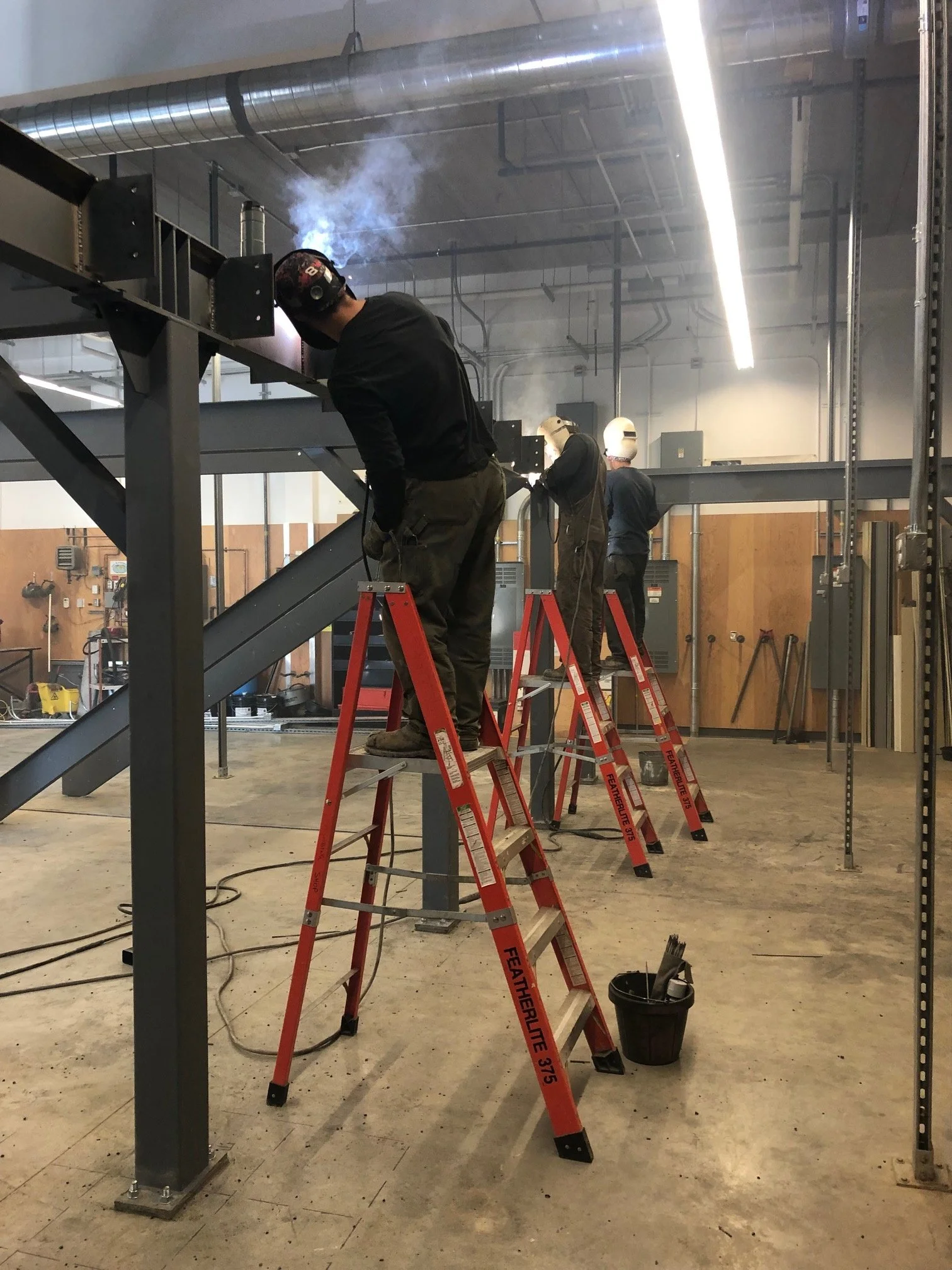Workers welding metal framework on orange ladders inside an industrial workshop, with smoke and industrial equipment visible.