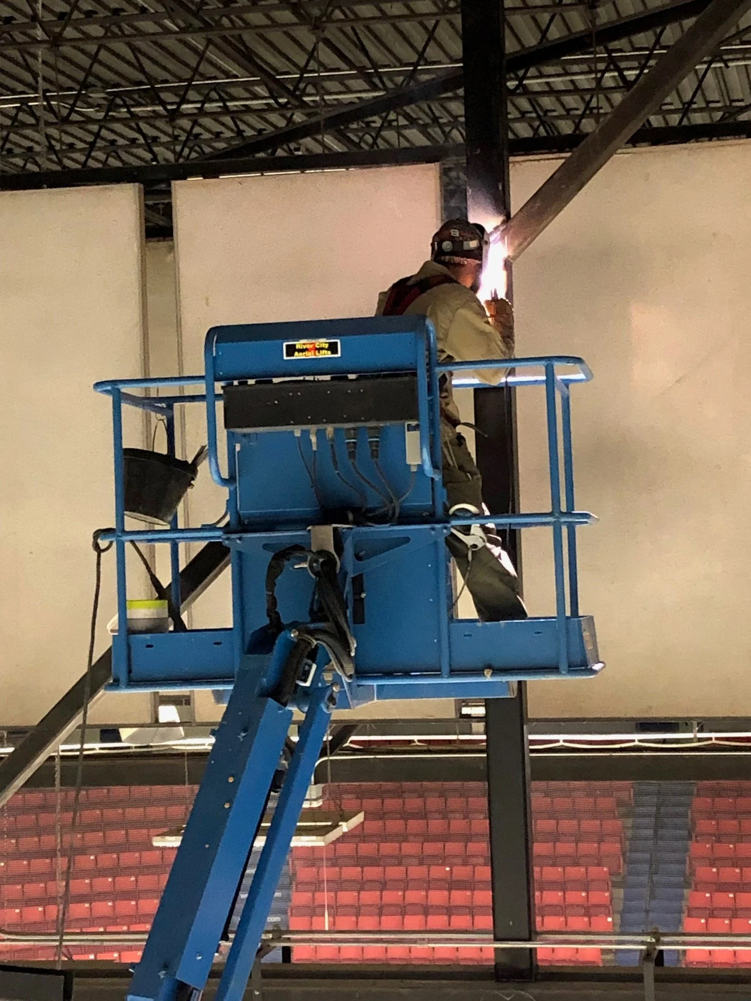 A worker welding metal beams while standing on a blue lift inside a building with a red and black seating area in the background.