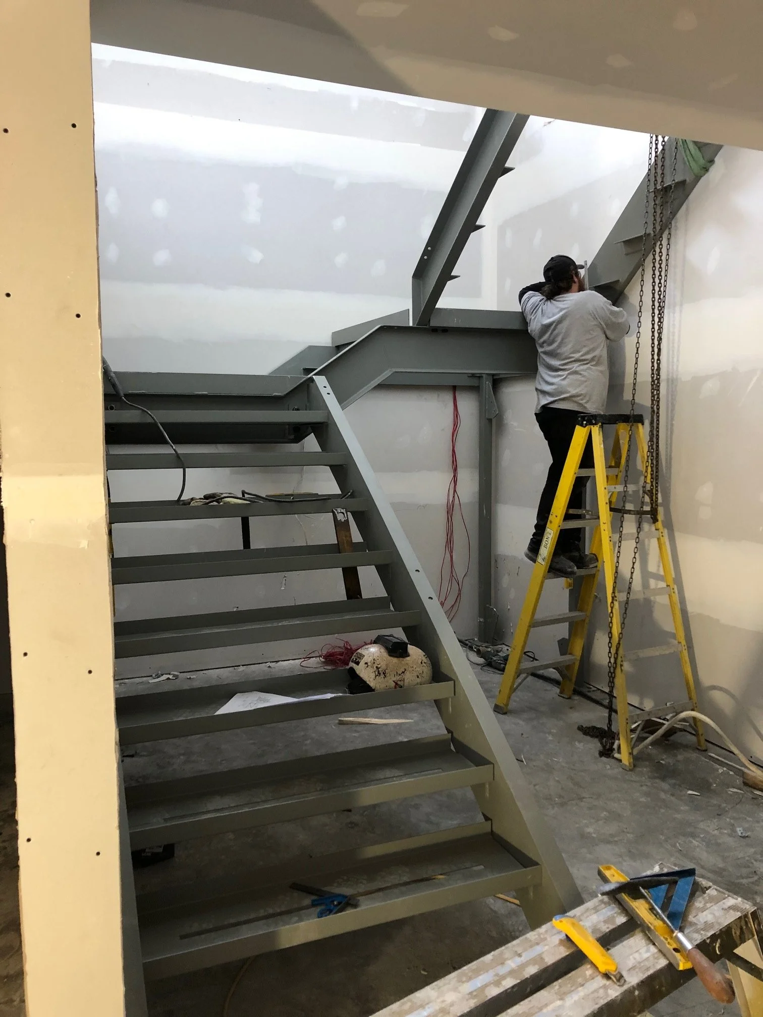 A person working on a metal staircase under construction, using a ladder in an indoor space with unfinished walls.