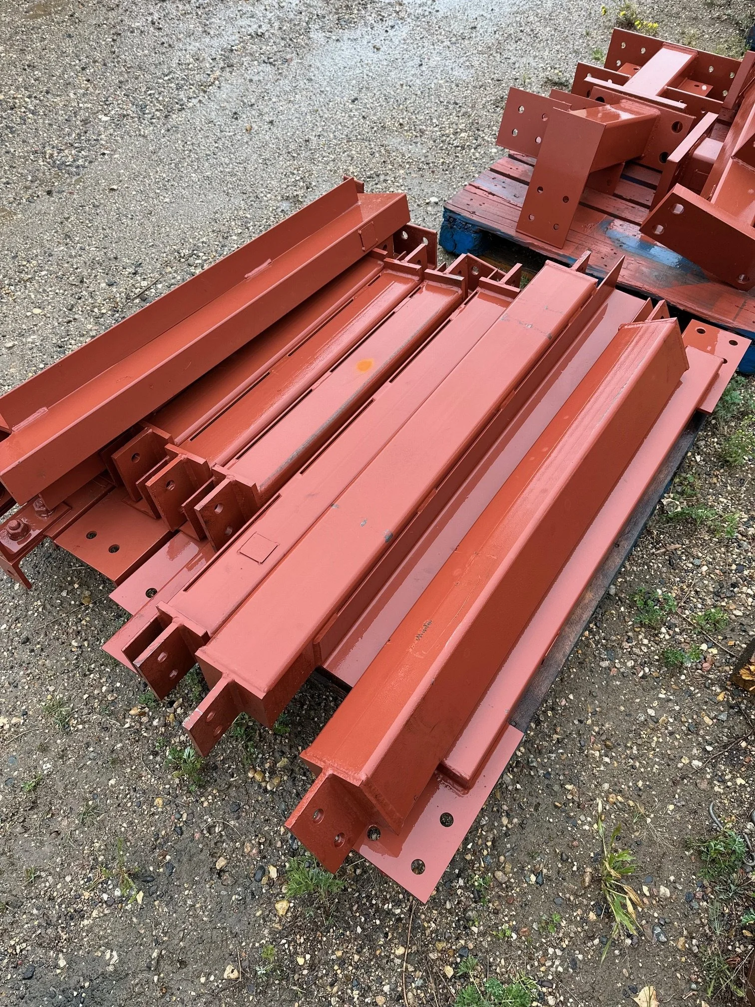 Stack of newly painted red steel beams and brackets on a wooden pallet outdoors on gravel.
