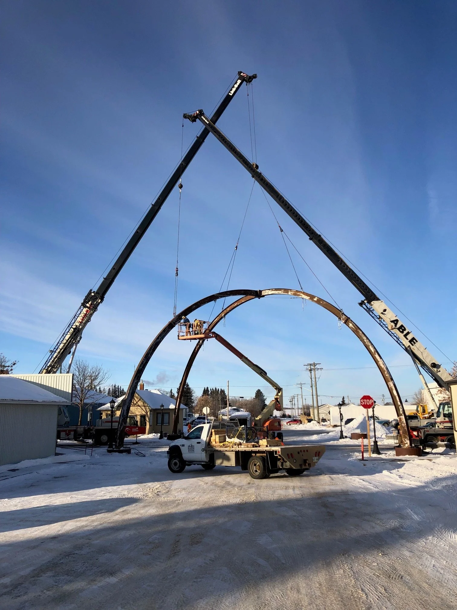 Workers use cranes to assemble large, metal arch structures outdoors on a snowy day in a residential area.