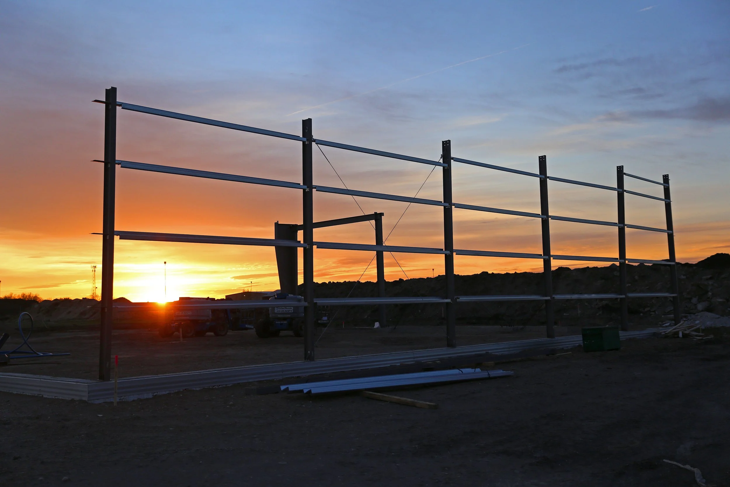 Steel framework of a building under construction during sunset, with construction vehicles and materials on the ground.