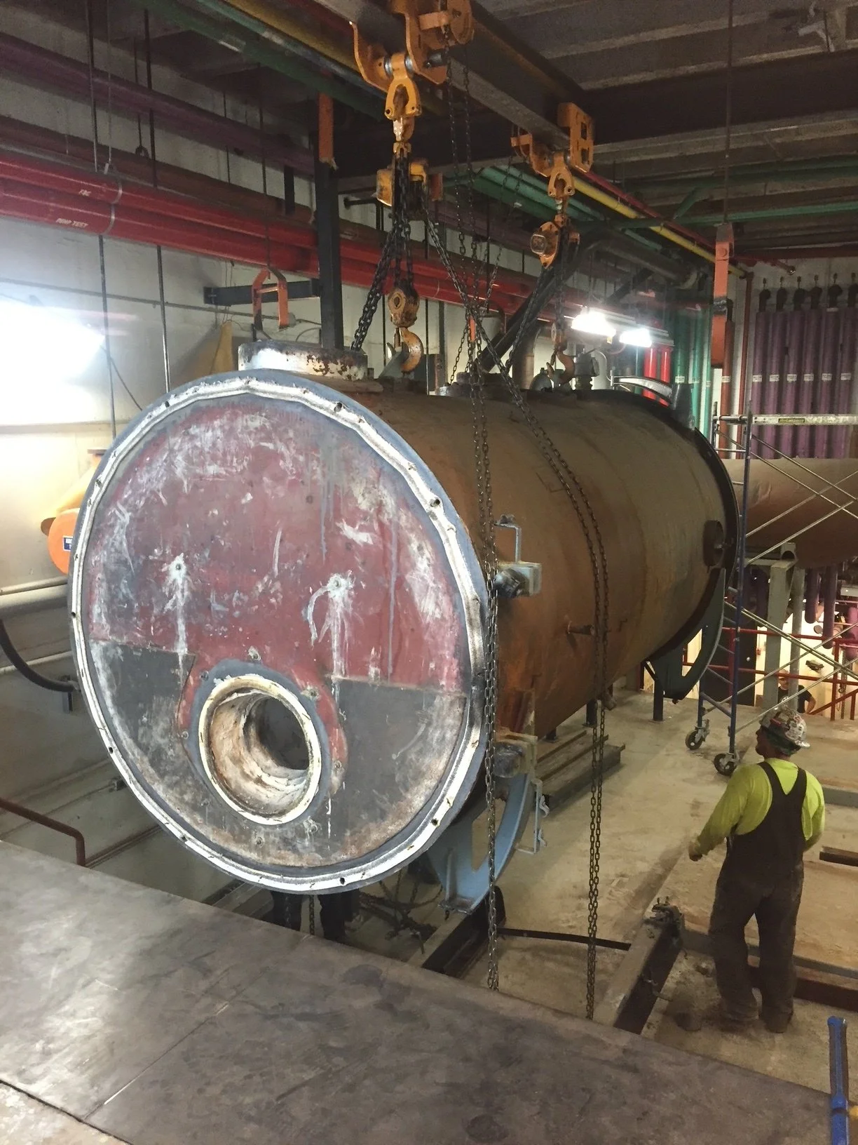 Large cylindrical industrial tank being installed or maintained, with a worker in safety gear nearby, inside a factory or plant with pipes and scaffolding.