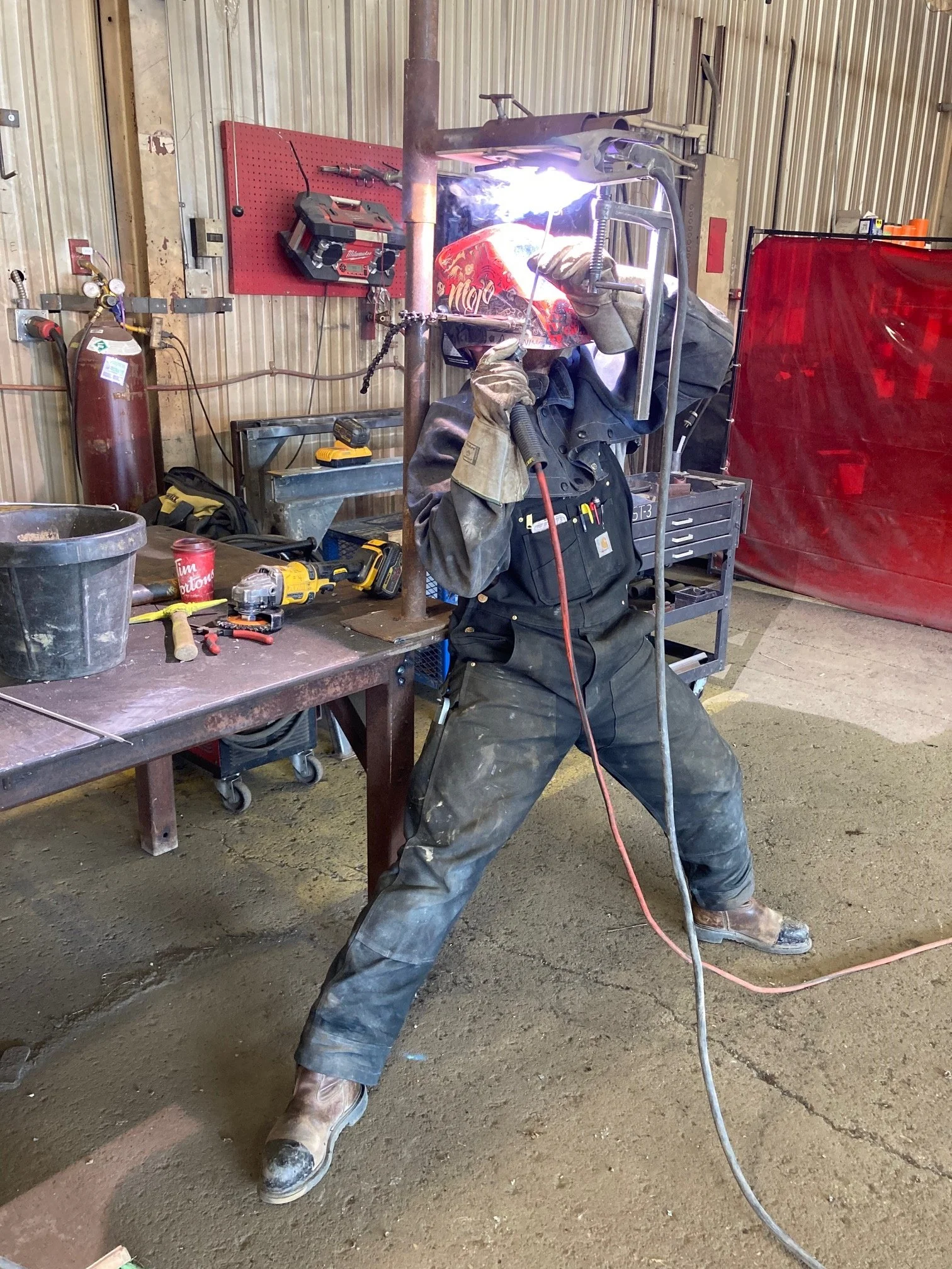 A person welding inside a workshop, wearing protective gear, including a welding helmet and gloves, with welding sparks visible.