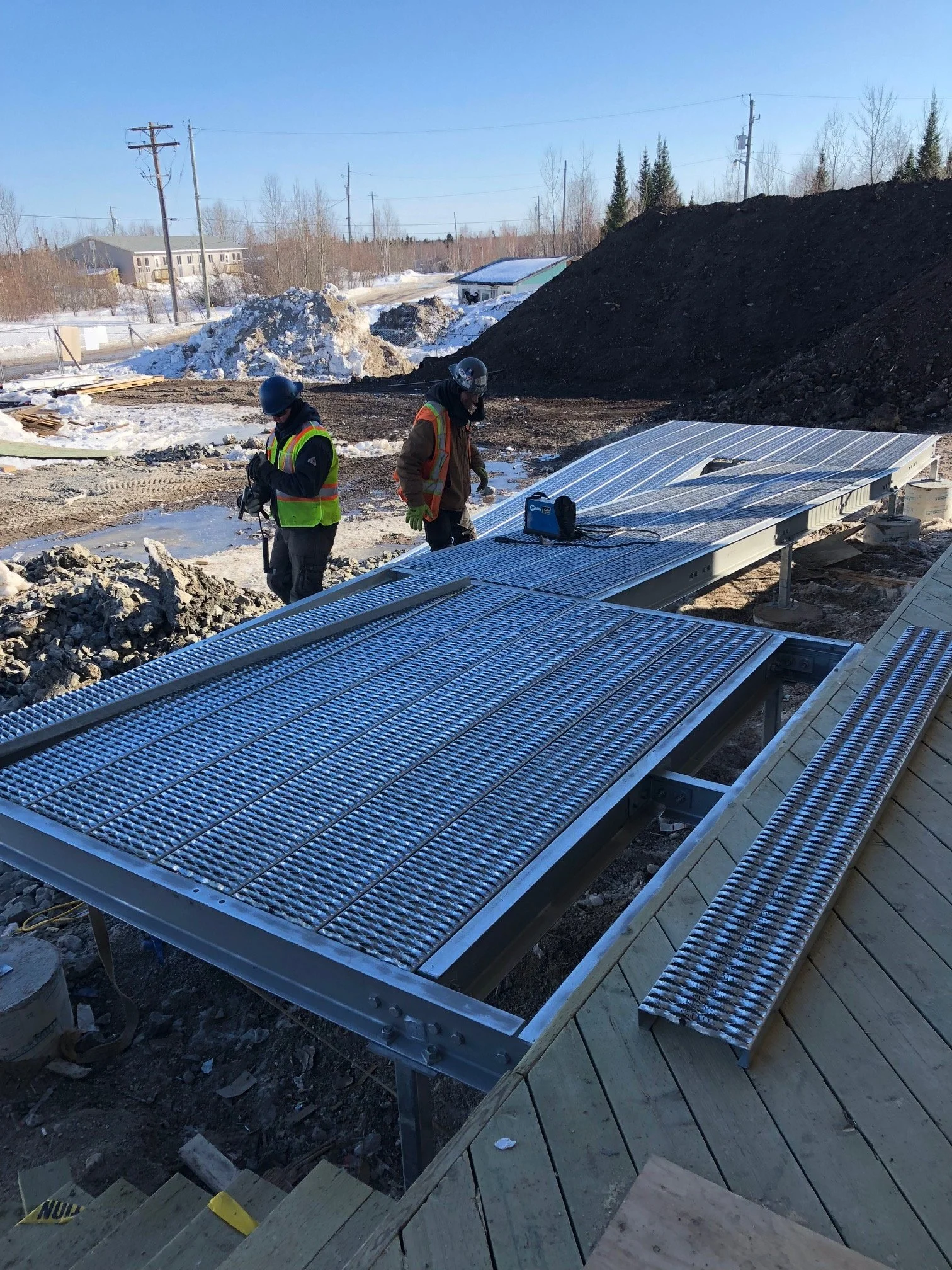 Construction workers installing solar panels on a roof outdoors on a building under construction with piles of snow and dirt nearby.
