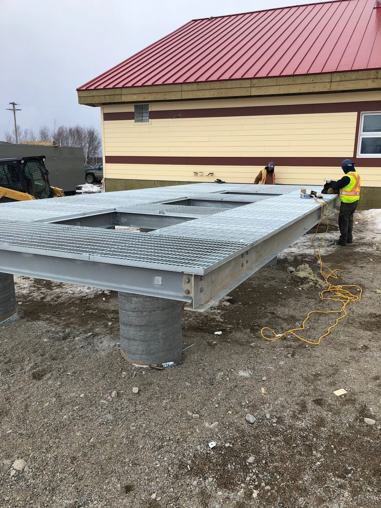 Construction workers installing a large metal platform or deck outside a building with beige and brown siding and a red metal roof, in a construction site with exposed dirt and gravel.