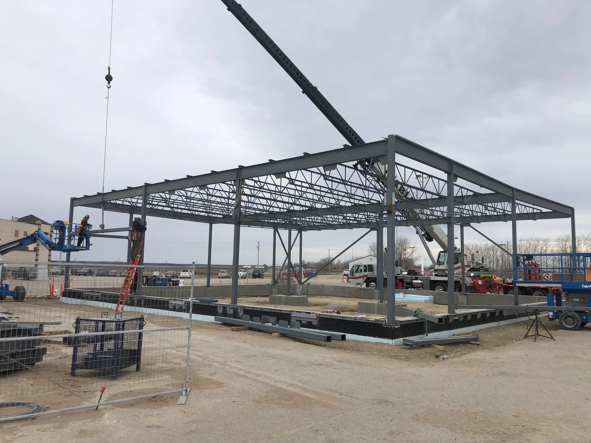 Construction workers assembling a steel framework for a building with cranes and equipment on a dirt lot.