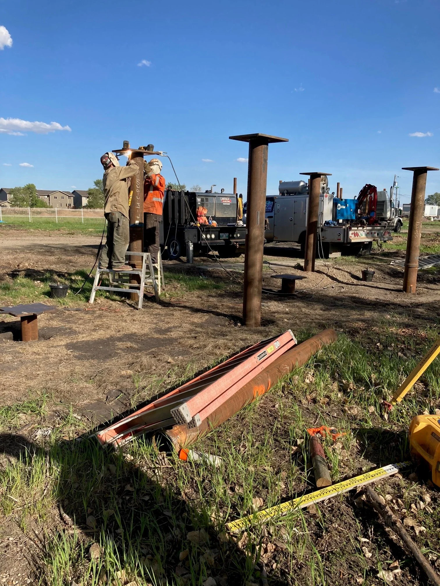 Two workers are installing or repairing utility poles at a construction site with trucks and equipment in the background, under a clear blue sky.