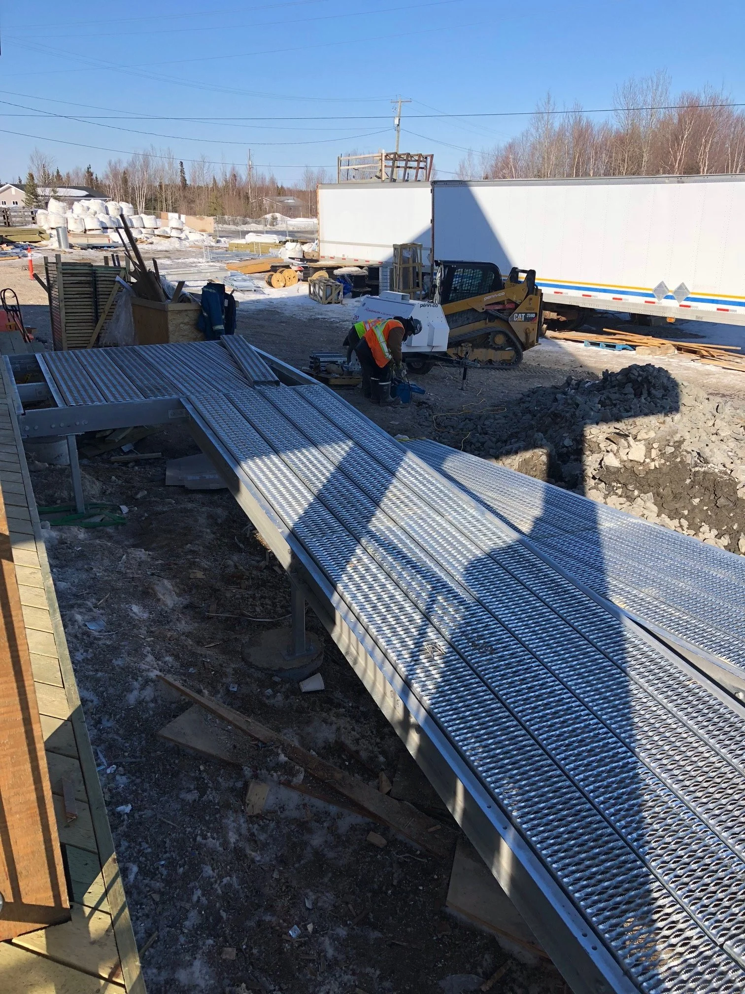 Construction site with worker operating machinery near a partially installed metal ramp, with trucks and construction materials in the background under a clear blue sky.