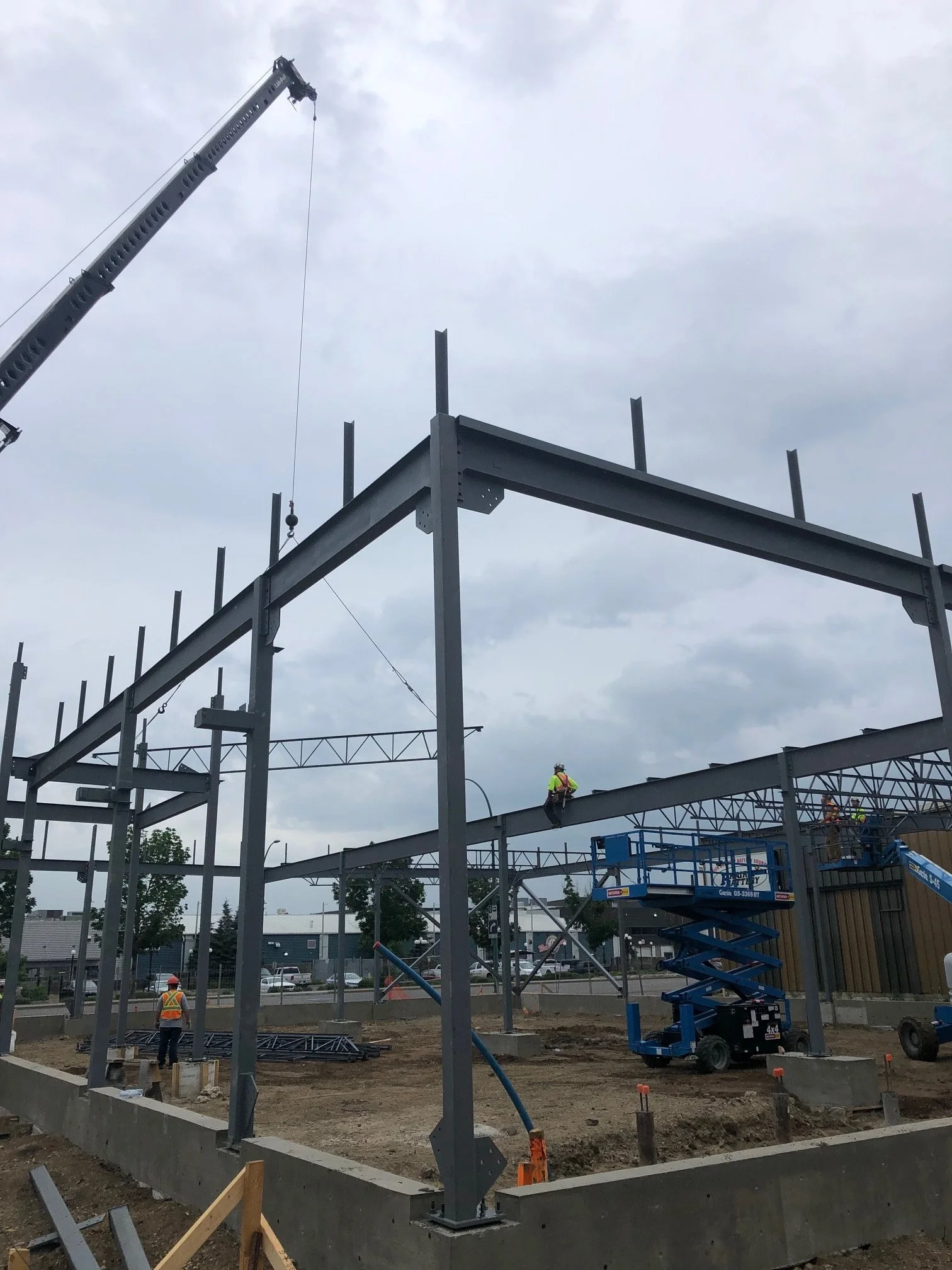 Construction workers building a steel frame structure with a crane and scissor lift on a cloudy day.
