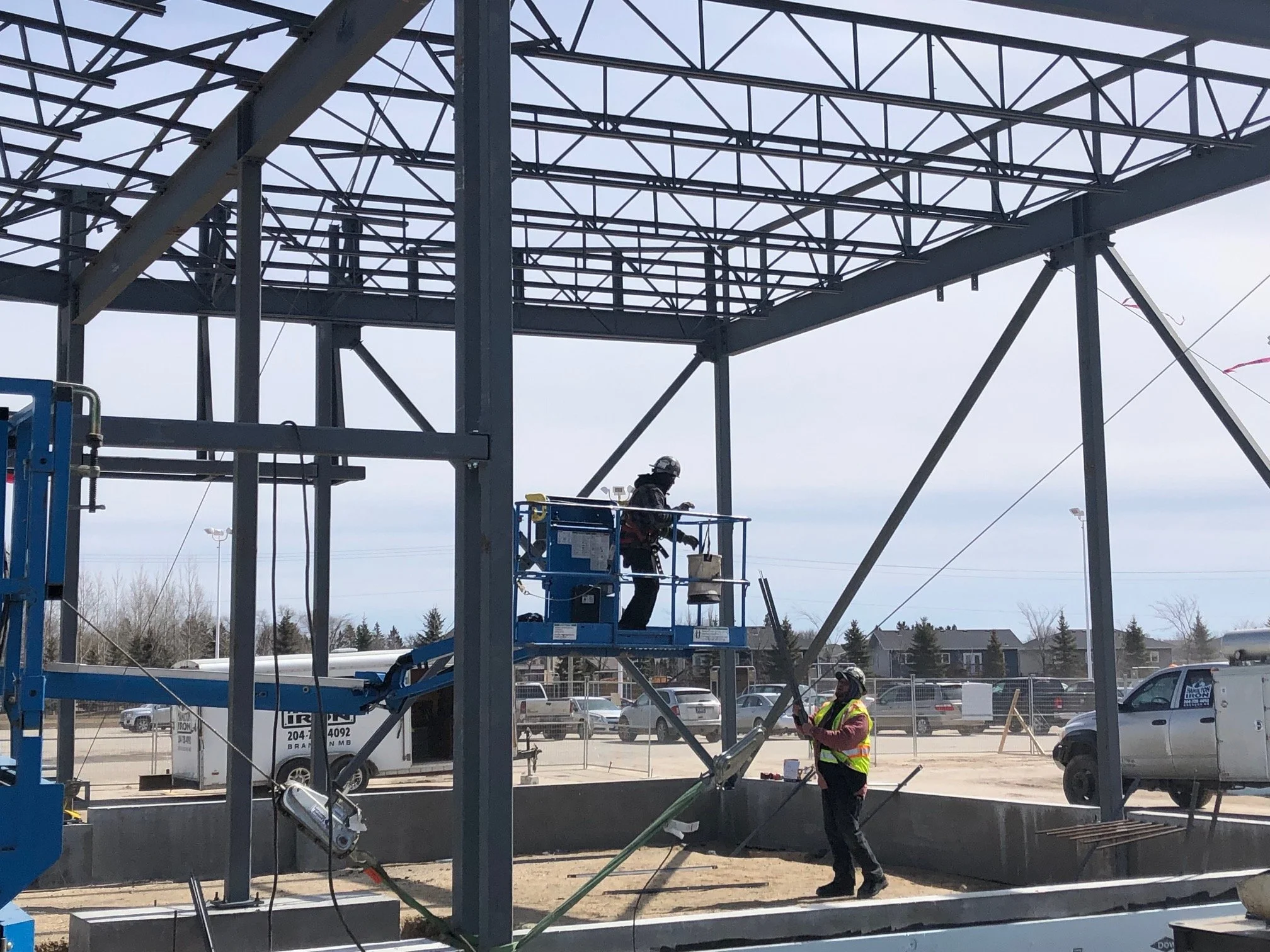 Two construction workers are building a steel frame structure outdoors, with one worker on a raised platform operating a power tool and the other worker standing below holding a support pole, in a parking lot area under a partly cloudy sky.