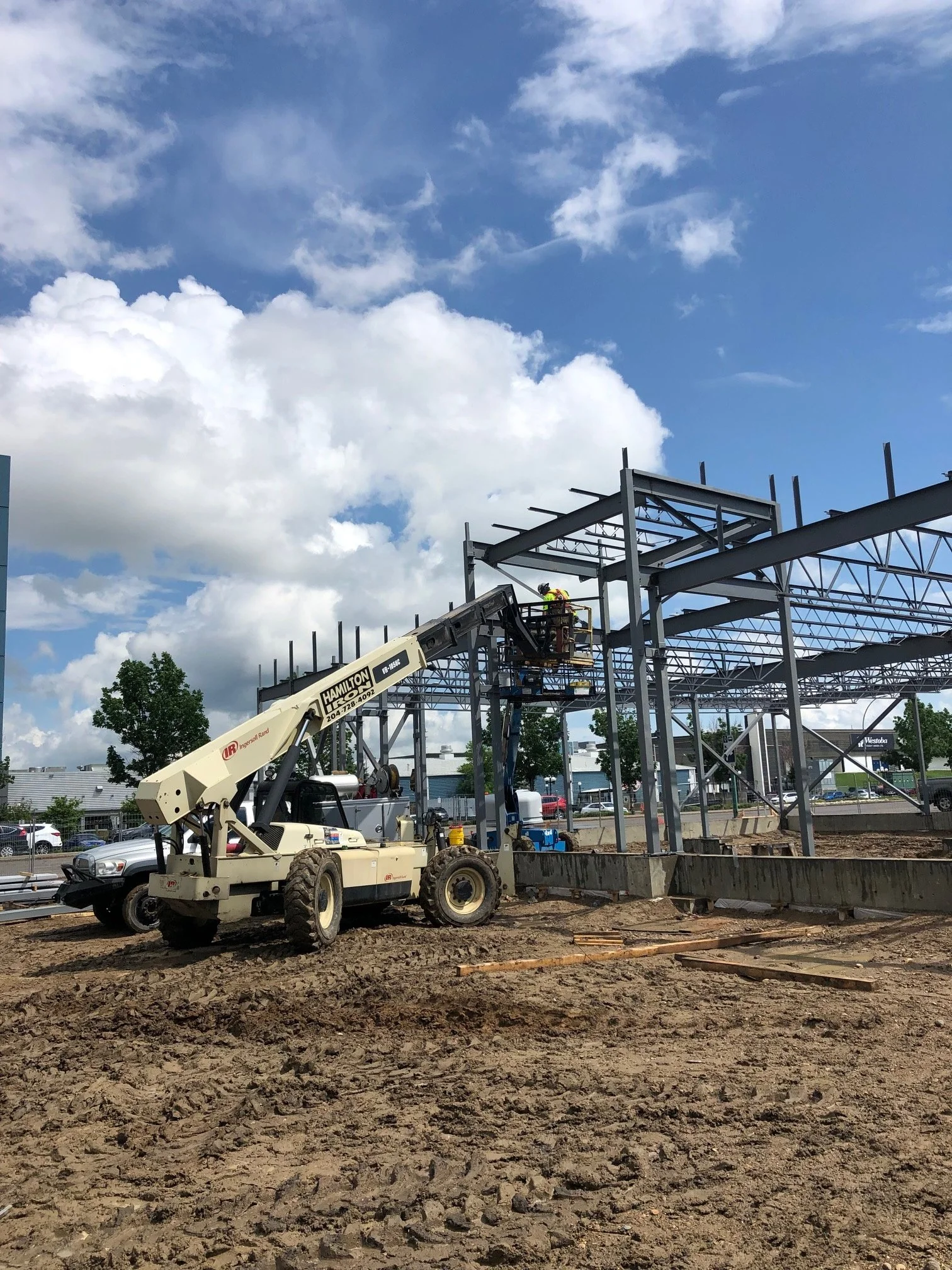 Construction workers working on steel framework at a building site, with a crane and construction tools, under a partly cloudy sky.