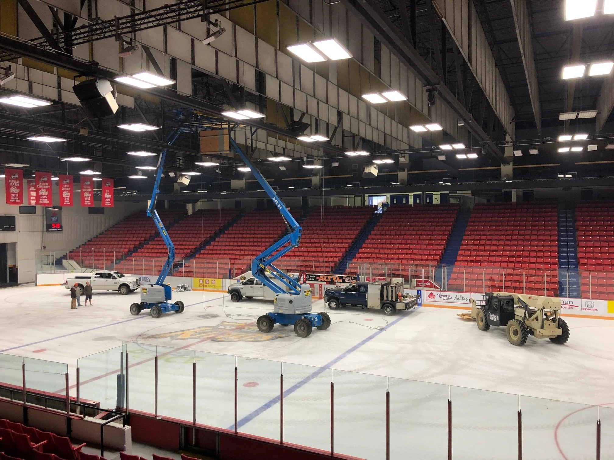 Empty indoor ice hockey rink with several construction vehicles and trucks, two workers standing on the ice, and red seats surrounding the rink.