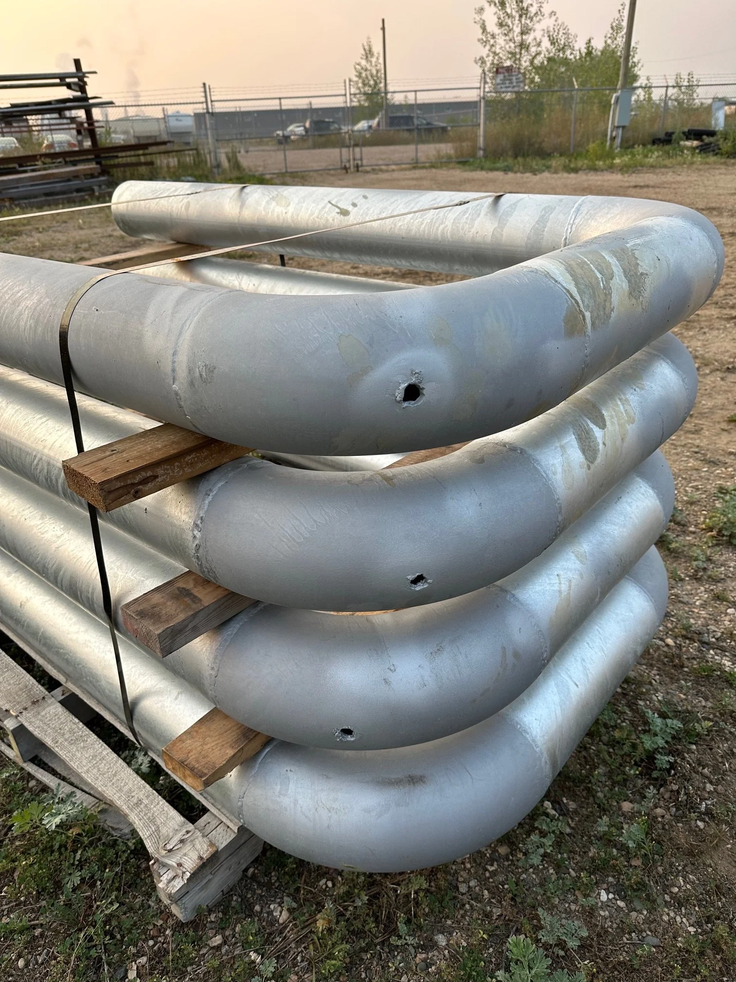 Stacked large metal pipes with holes, secured with wooden blocks and black straps, outdoors on a dirt surface with a fence and industrial area in the background.