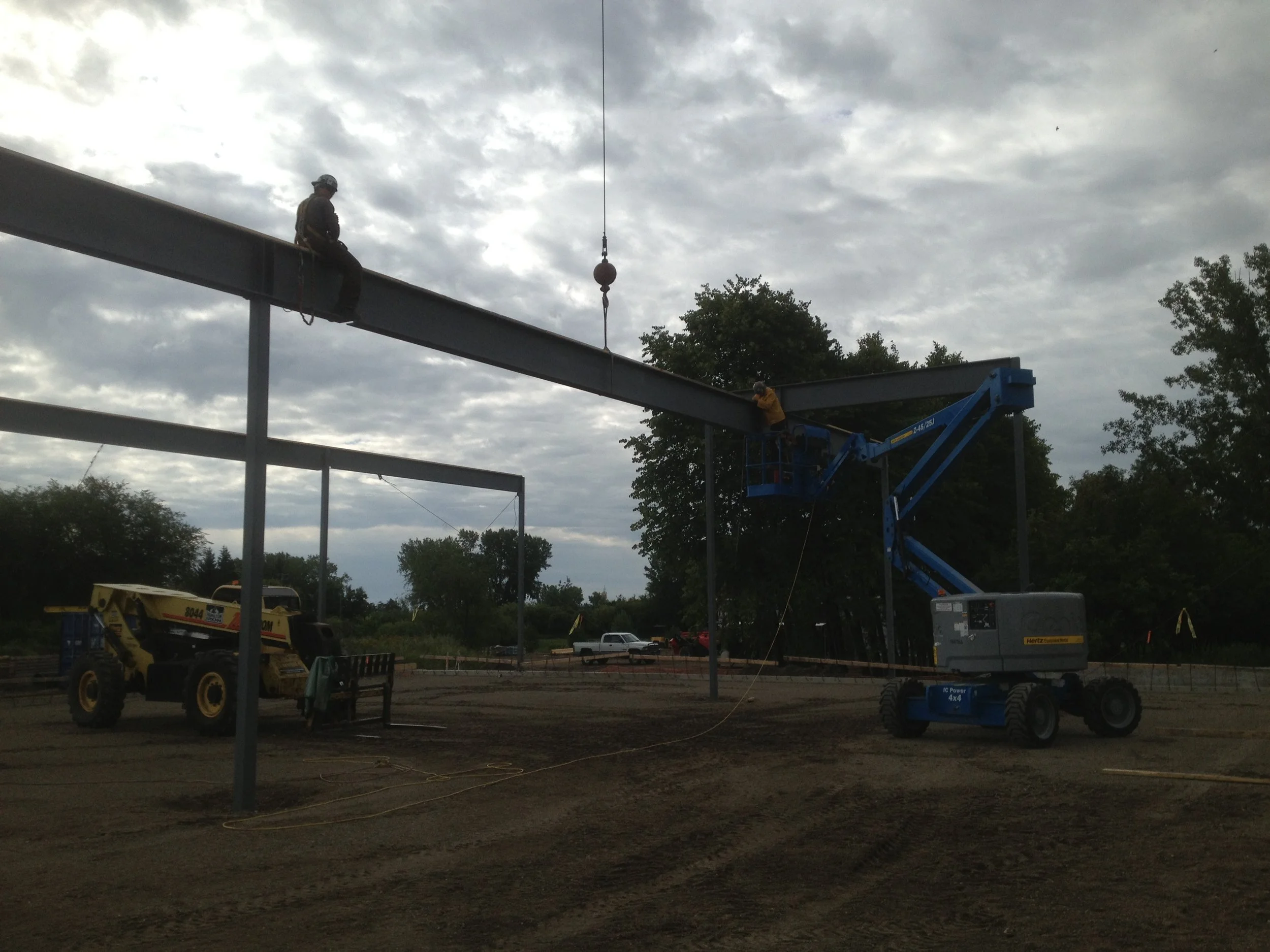 Construction site with steel beams being assembled, two workers on a blue lift, one worker sitting on a steel beam, over a dirt ground with cloudy sky in the background.