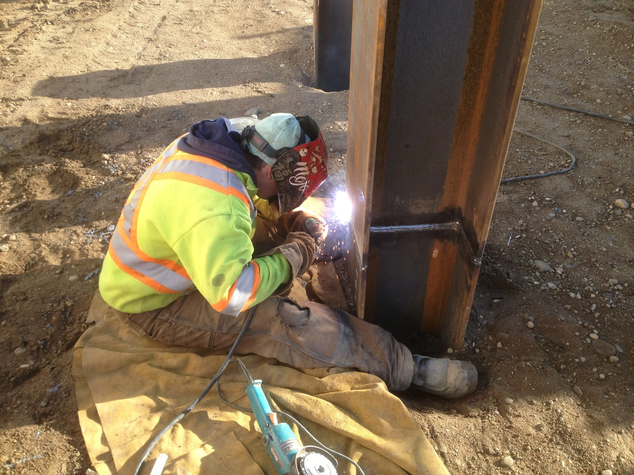 A construction worker welding metal beams at a construction site on a dirt surface.