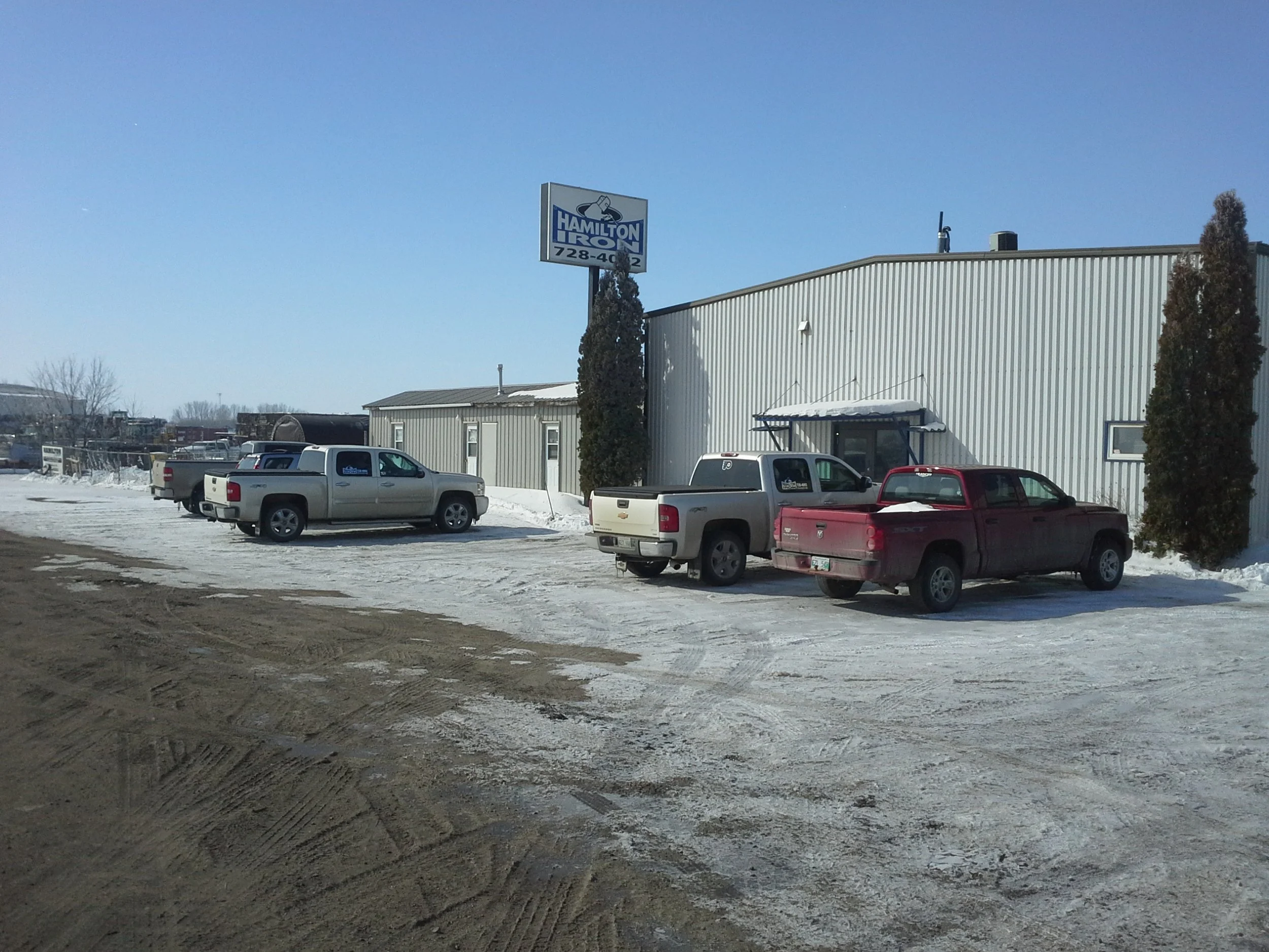 Parking lot with snow and four pickup trucks parked in front of a metal building with a sign reading 'HAMILTON IRON' and a phone number. Clear blue sky.