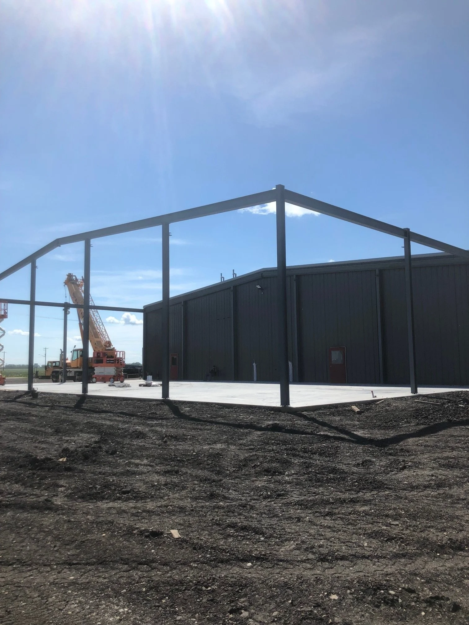 Construction site with a partially built metal structure, black building, and a crane in the background under a sunny sky