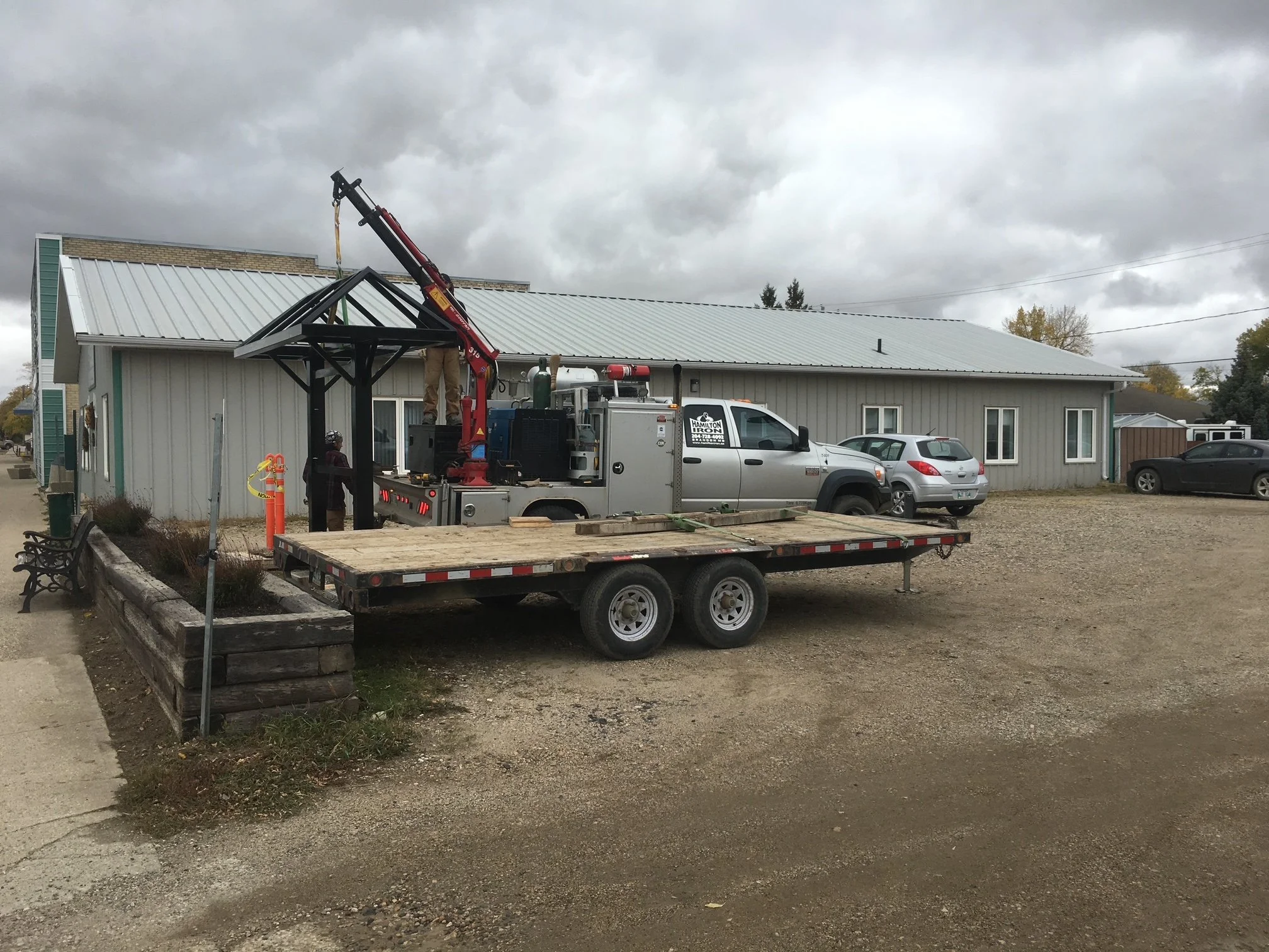People installing a small glass roof on a black metal structure outside a building, using a crane on a flatbed truck.