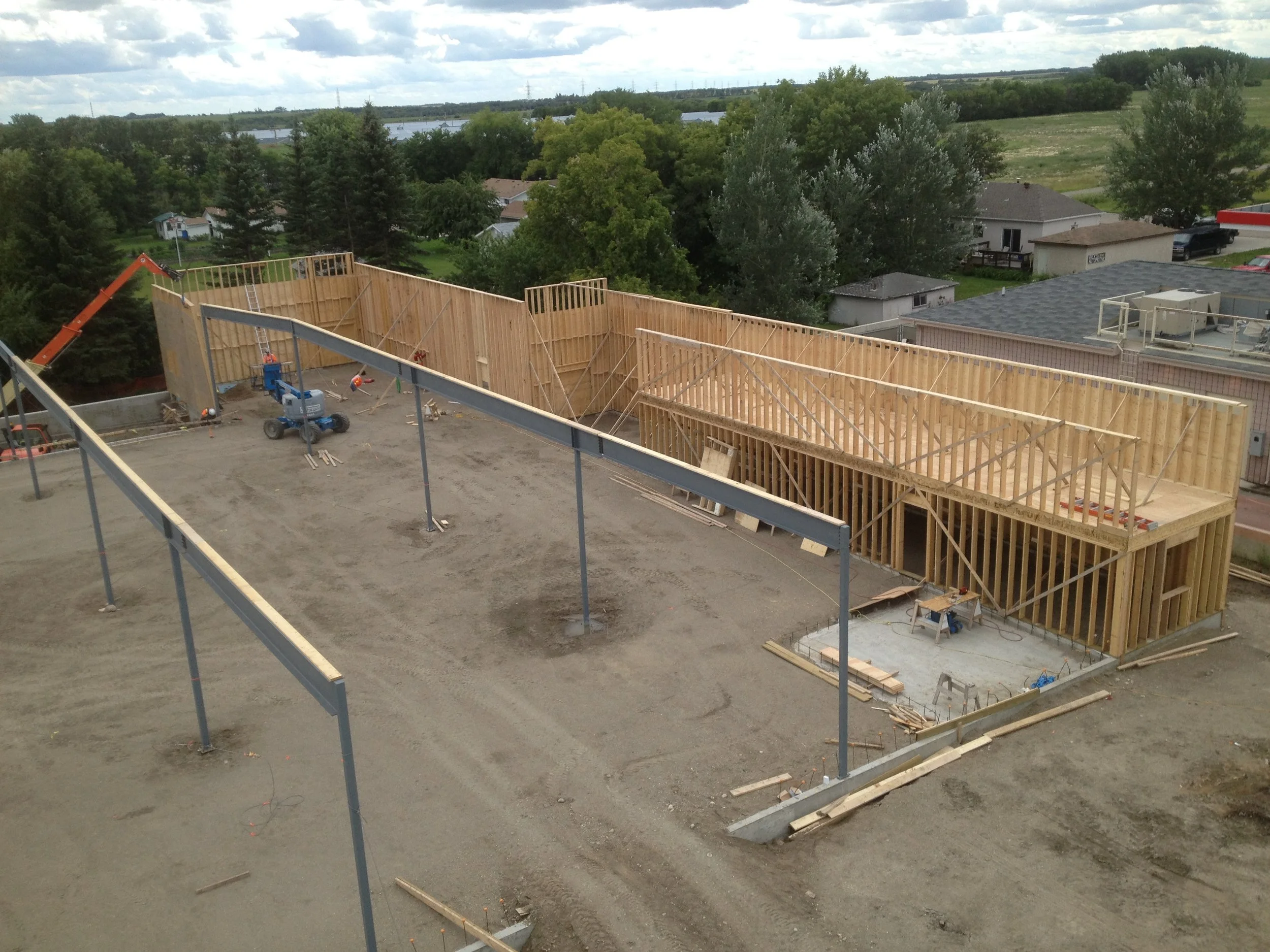 Construction site with wooden framing for a new building, partially built with support beams and tools on site, in a residential area with trees and houses in the background.