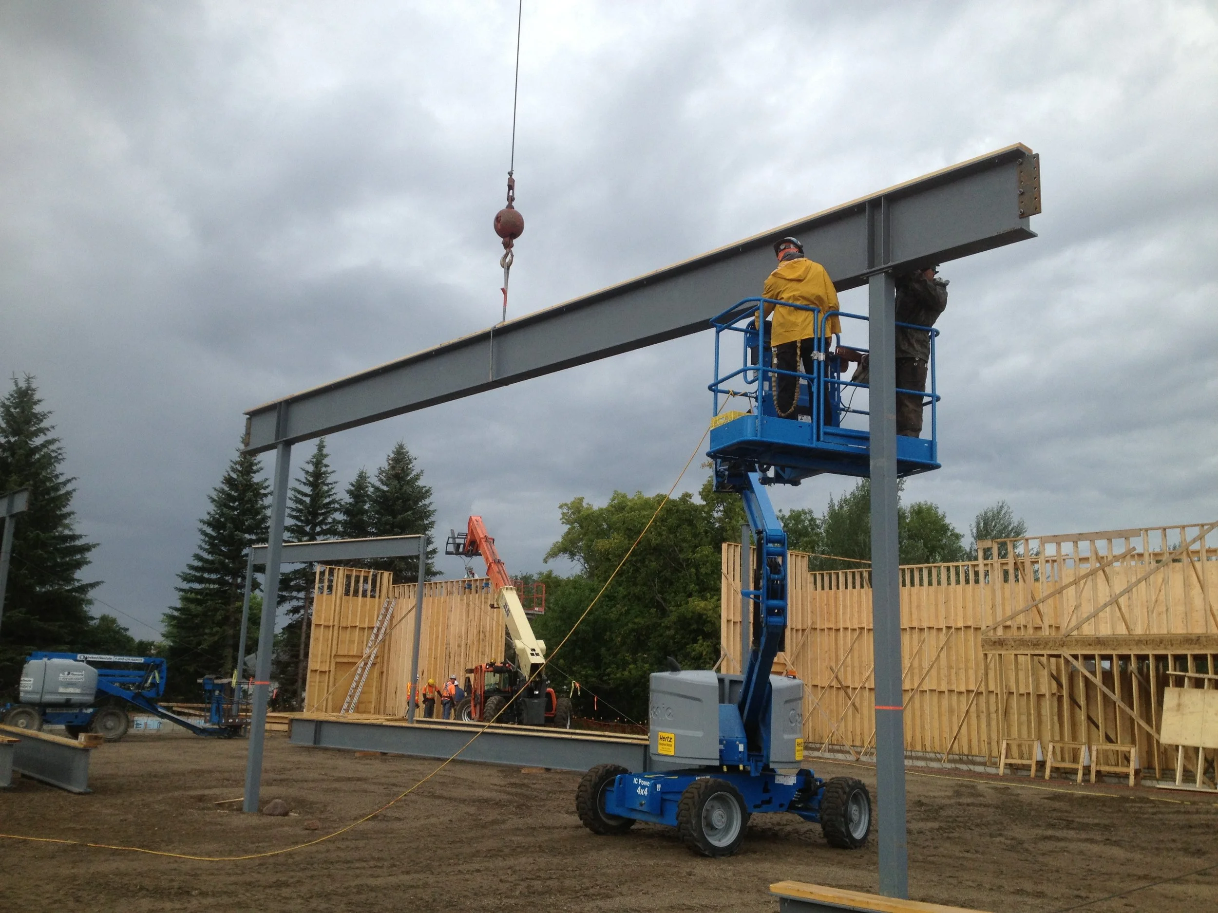 Two construction workers in a blue lift working on a steel beam at a construction site with wooden frame structures and construction equipment in the background.