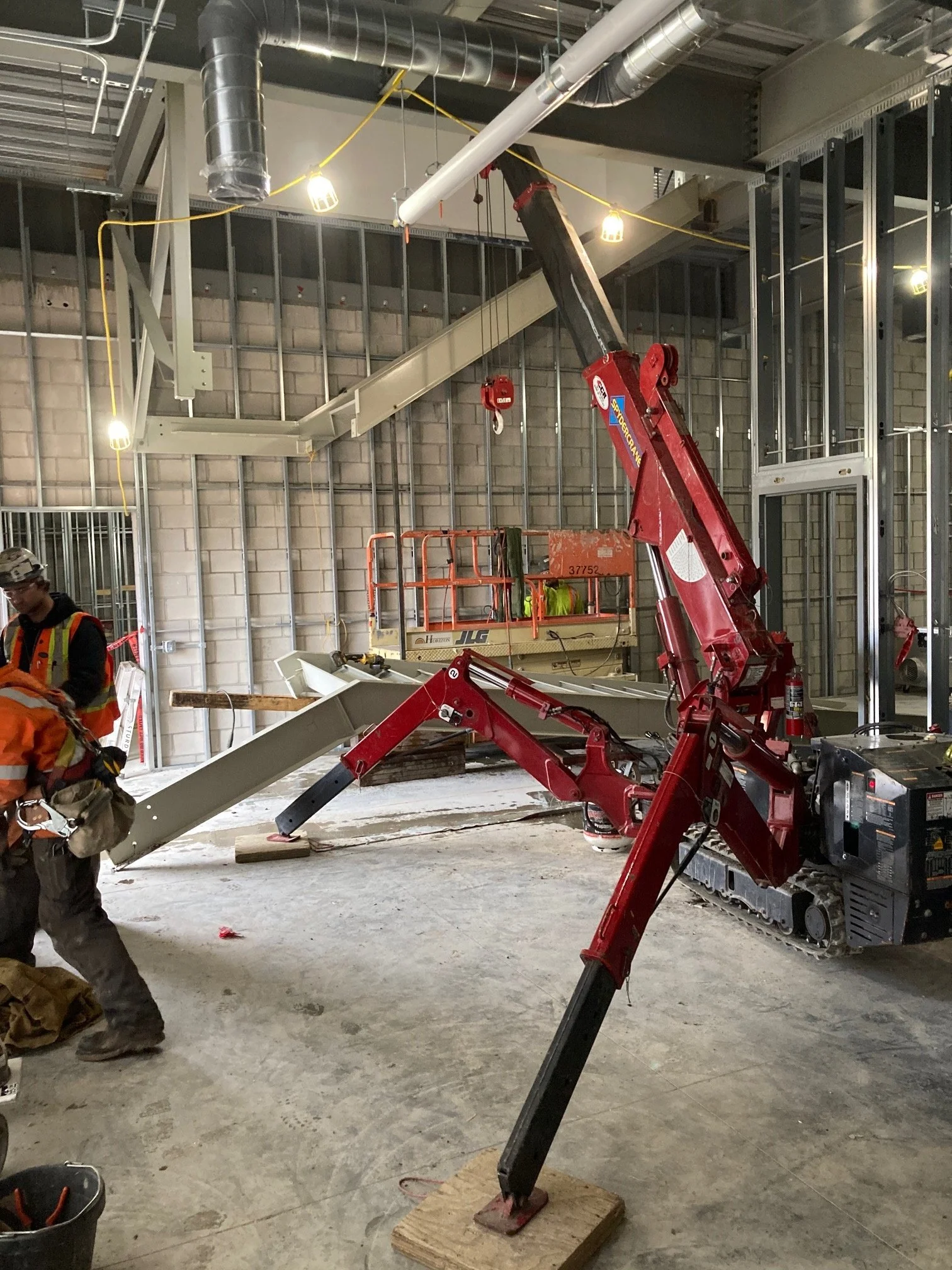 Construction workers are using a red telescopic boom lift to reach the ceiling of an unfinished building with exposed ductwork, pipes, and a metal framework.