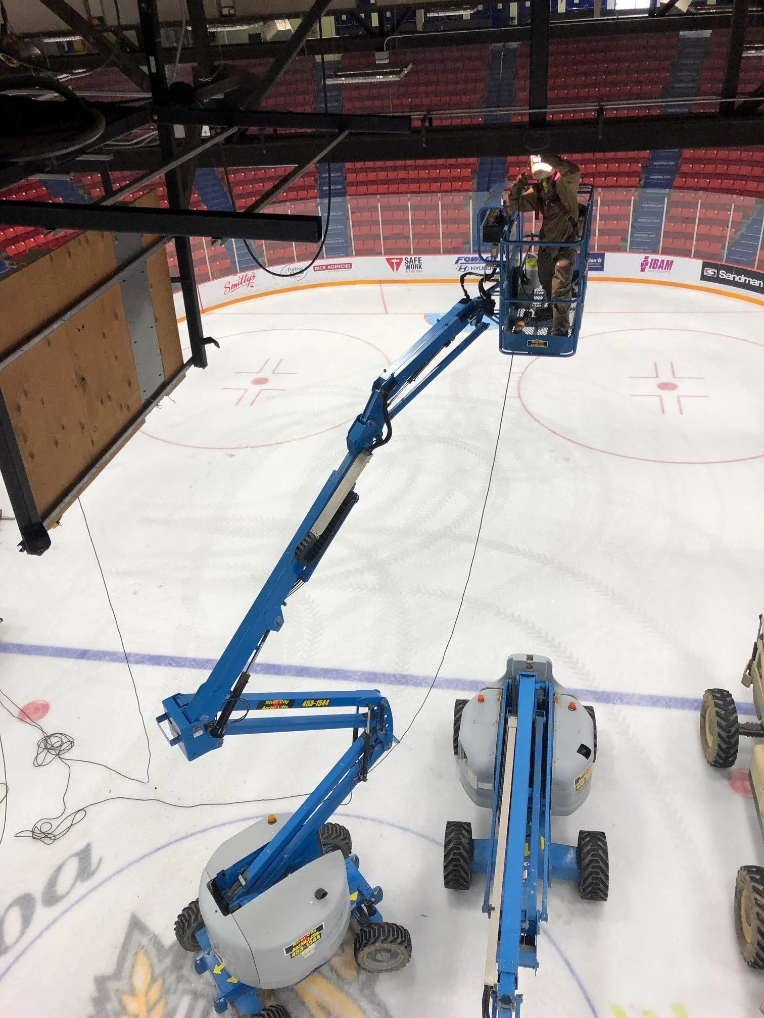 A worker on a lift cleaning or maintaining the ice rink inside an empty hockey arena.
