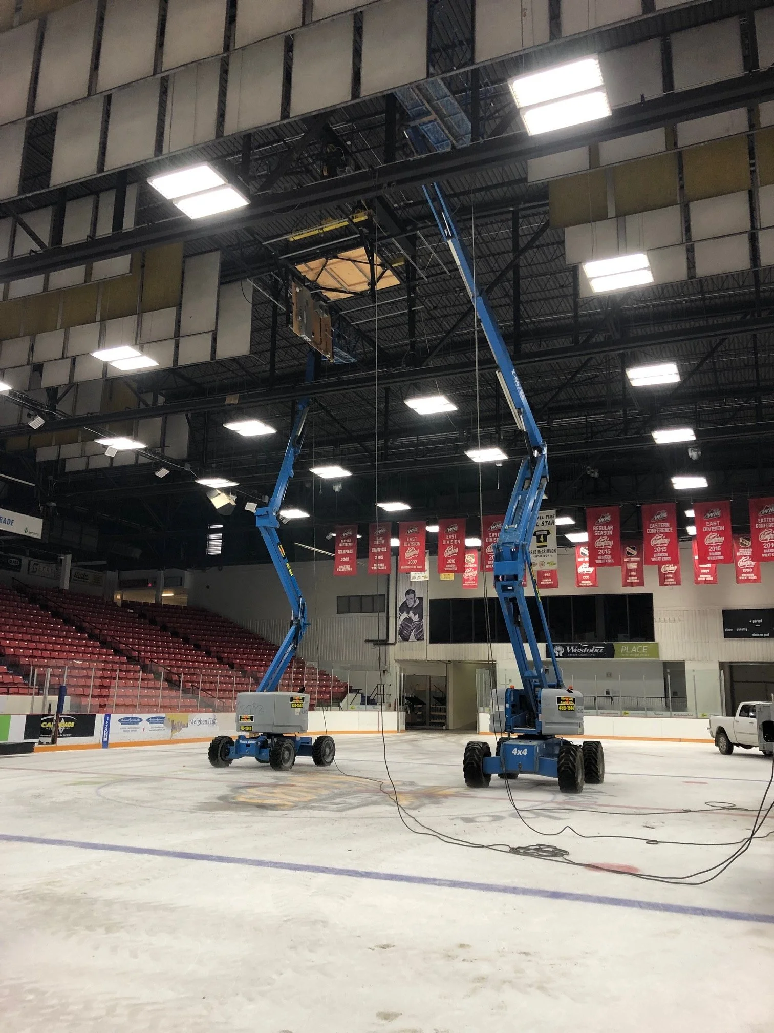 Two blue cherry pickers are on the ice rink, repairing or maintaining the ceiling above the ice. The rink is empty, with red seats and banners hanging from the ceiling.