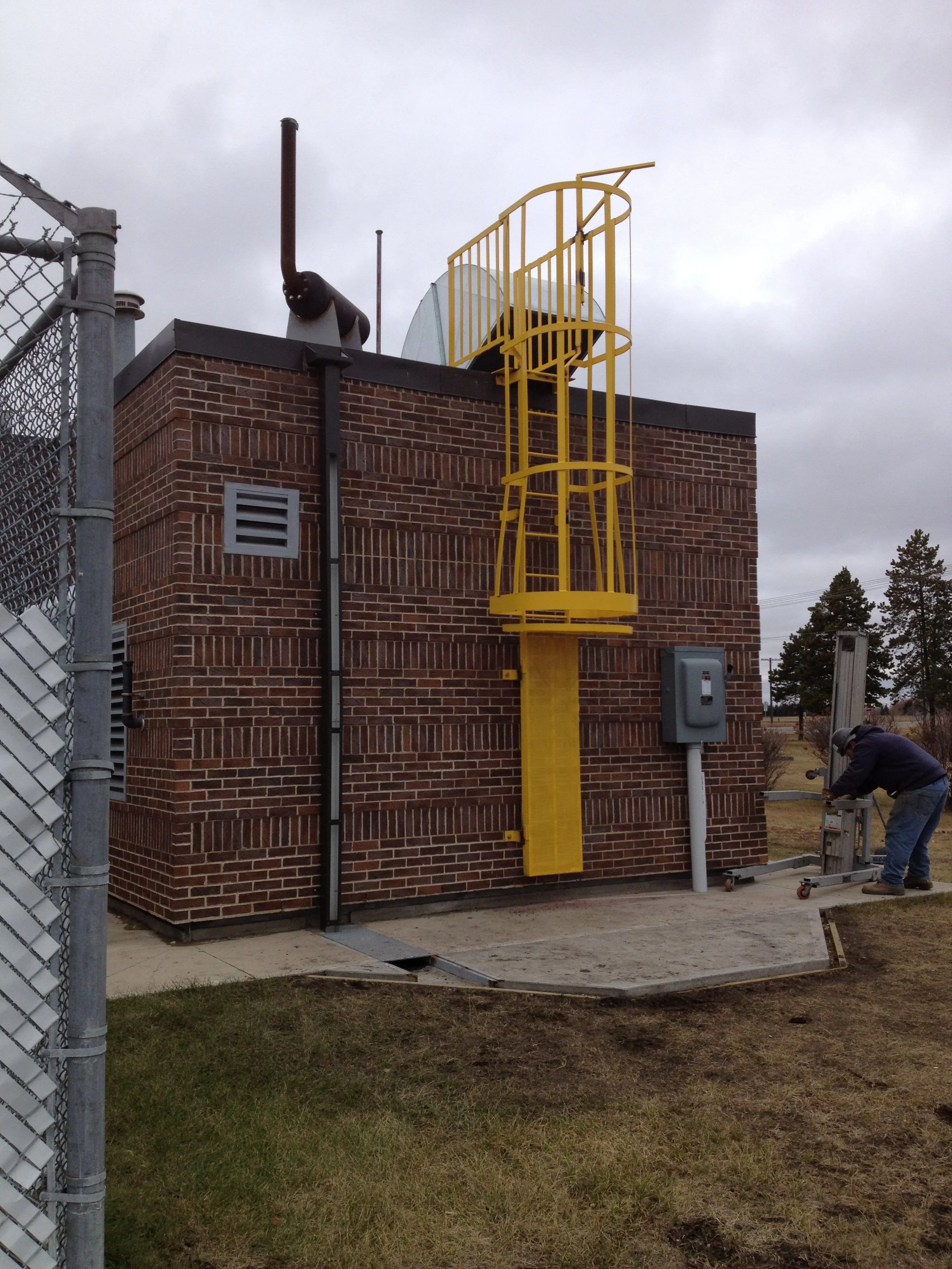 The side of a brick building with a yellow safety ladder attached to the wall, surrounded by construction tools and a worker working nearby under a cloudy sky.