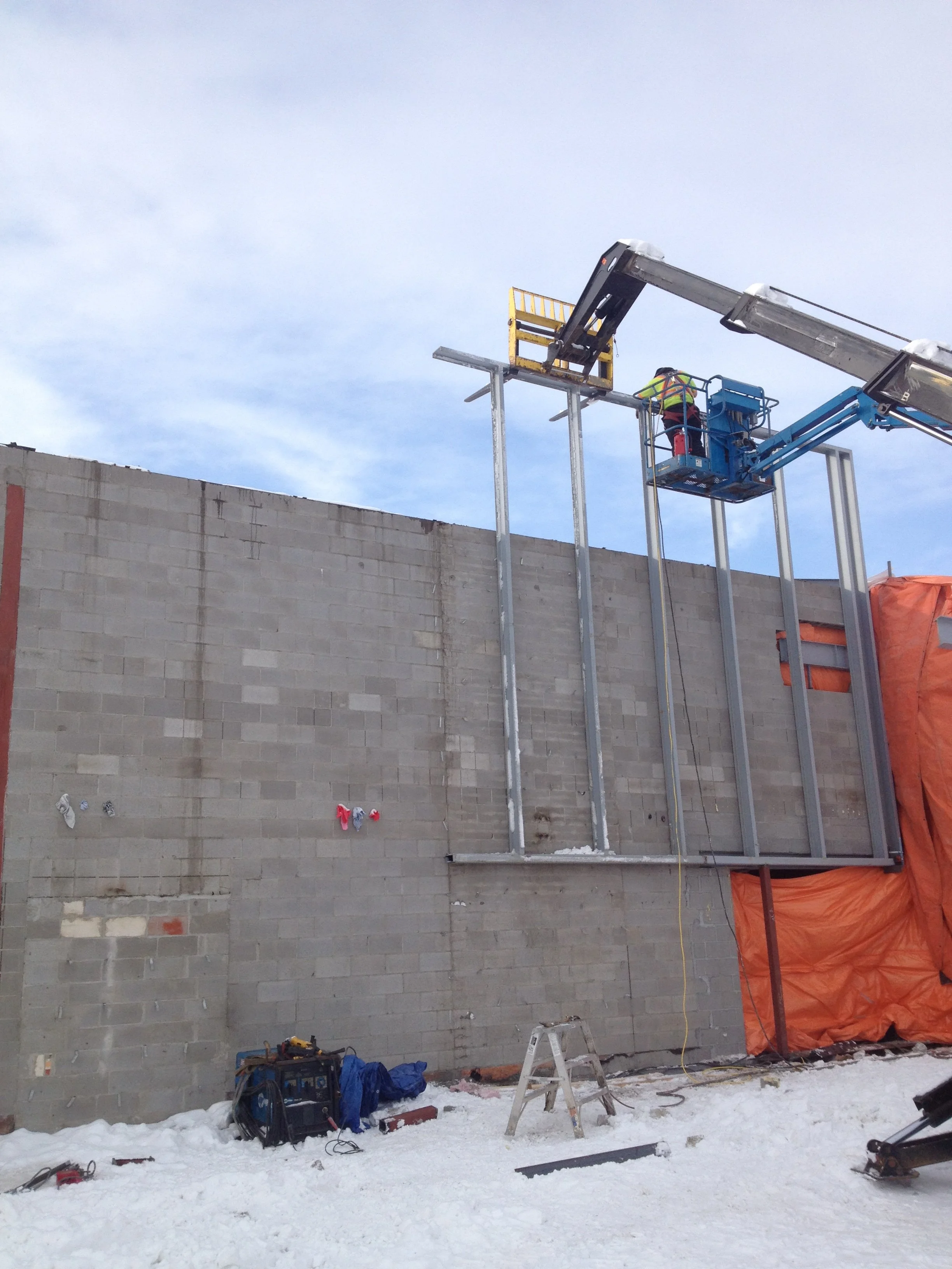 Construction workers installing steel framing on a building exterior, with snow on the ground and a cloudy sky.