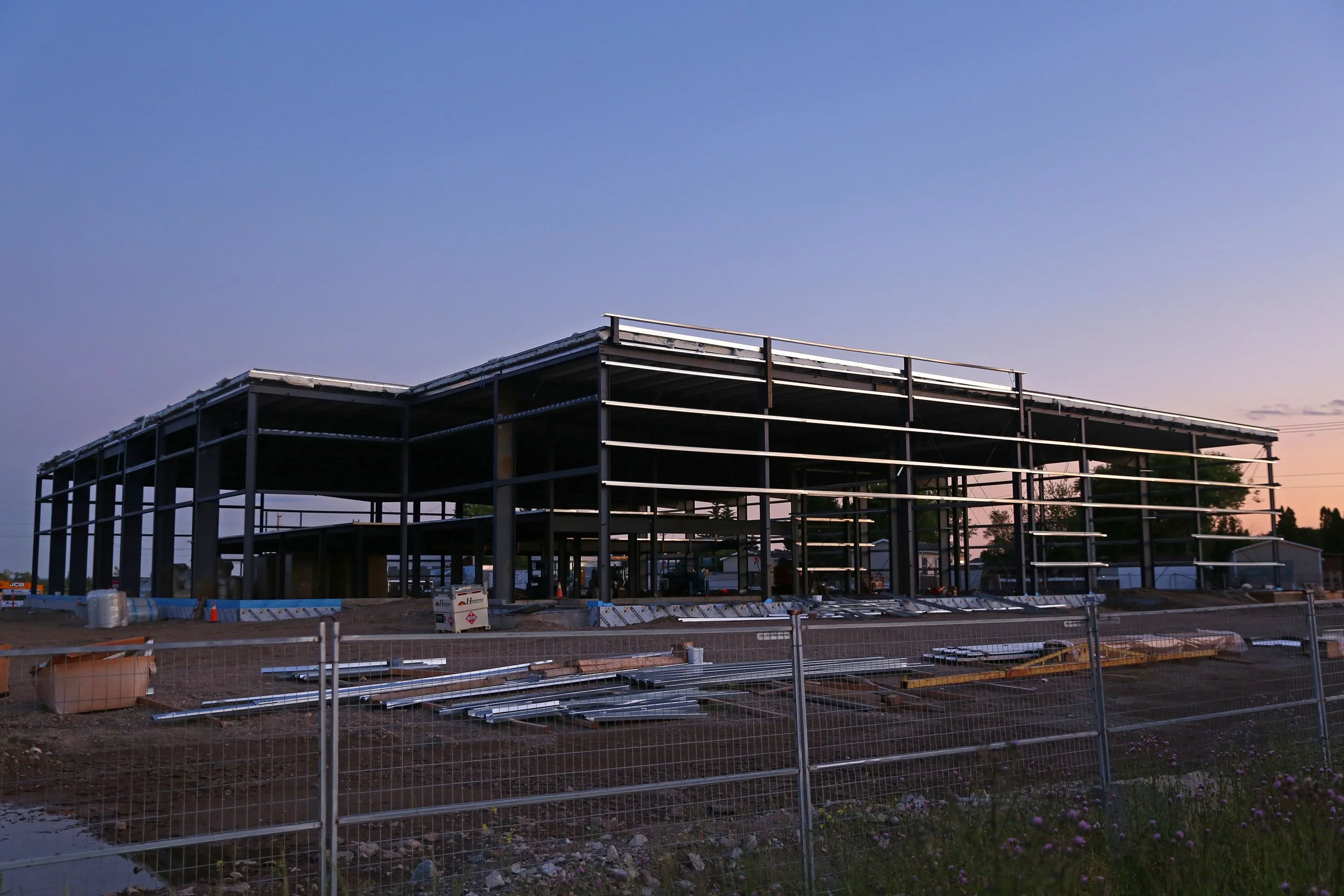 Construction site of a building framework with steel beams at dusk, with a fence in the foreground and trees in the background.