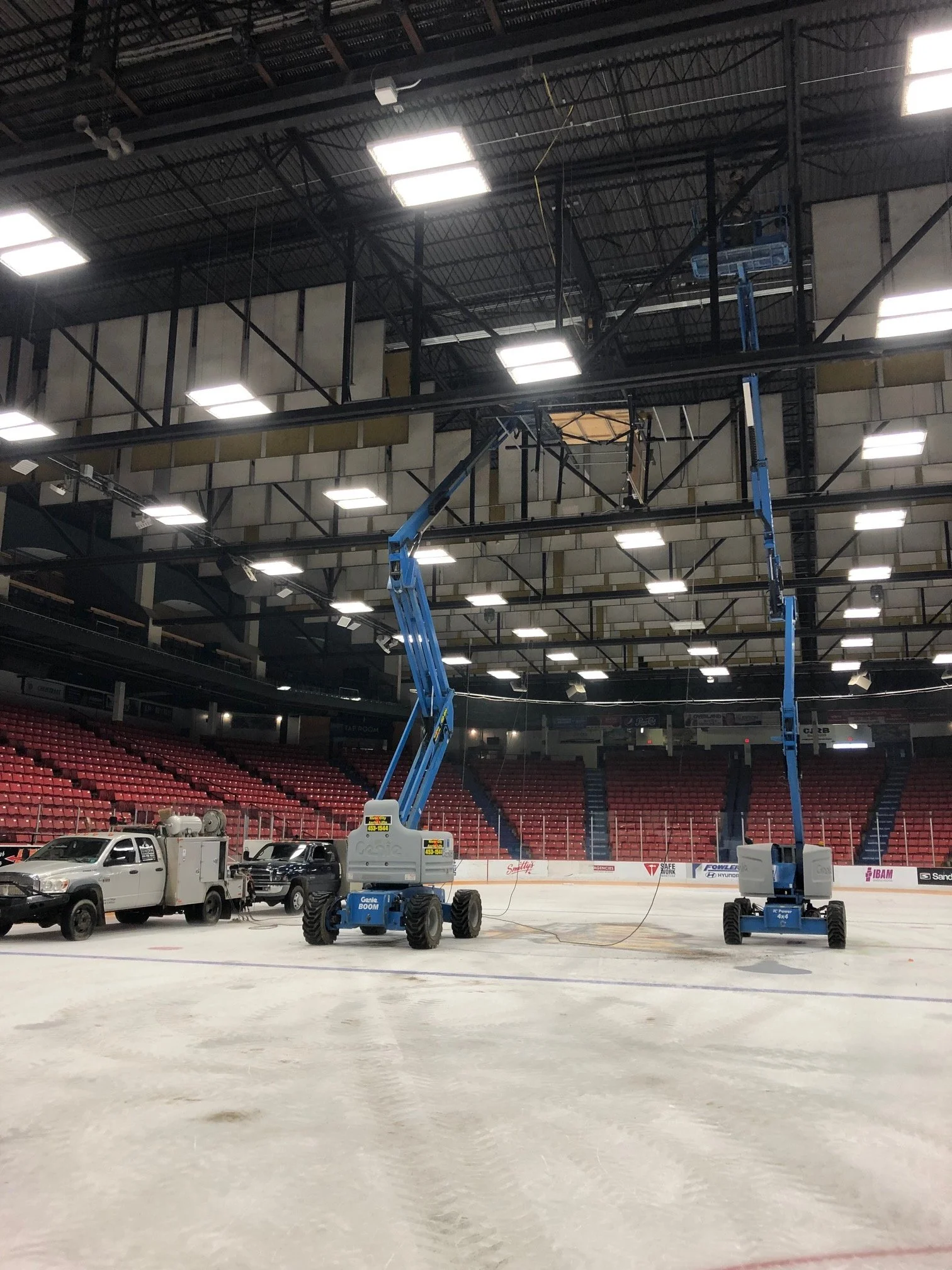 Inside an ice hockey arena under renovation, with two blue lift machines and a few parked vehicles, empty seating in the background, and bright ceiling lights.