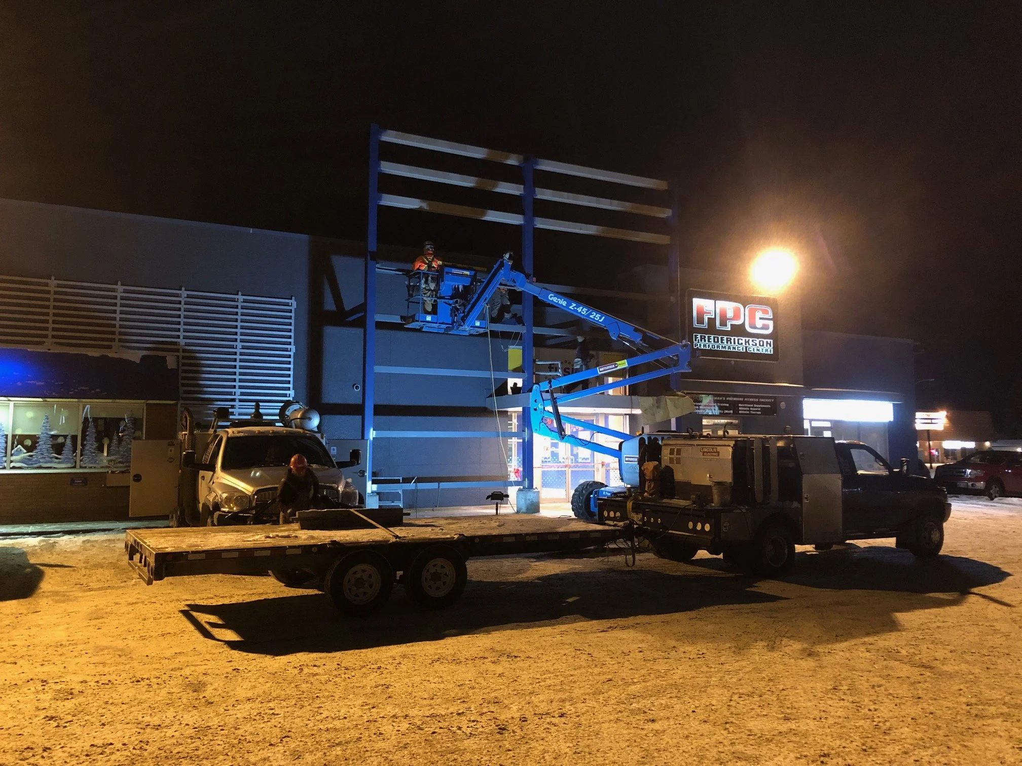 Workers on a blue cherry picker lift installing or repairing lights on the outside of a commercial building at night, with a tow truck nearby.