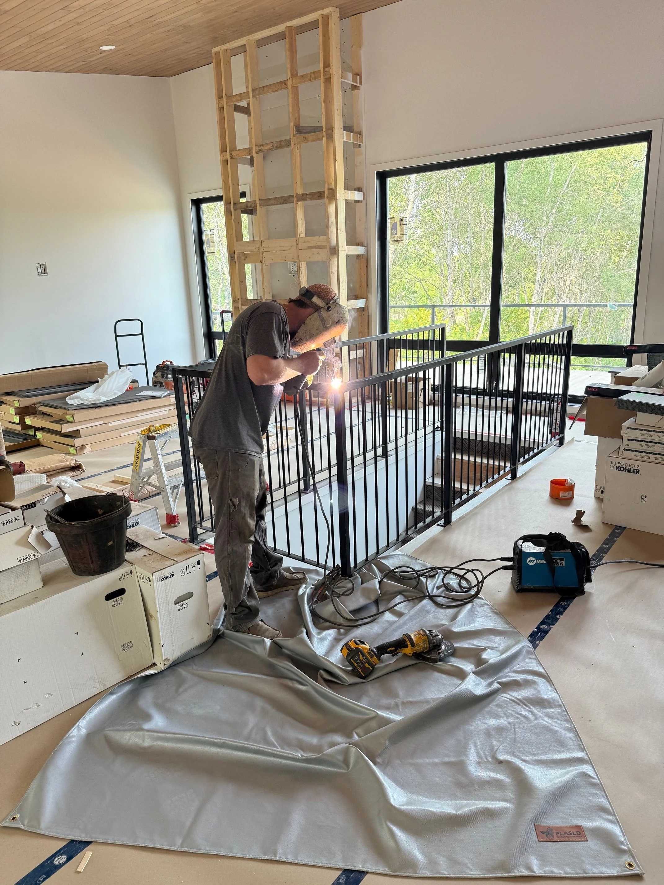 A man welding black metal railings inside a room under construction, with tools and construction materials scattered around.