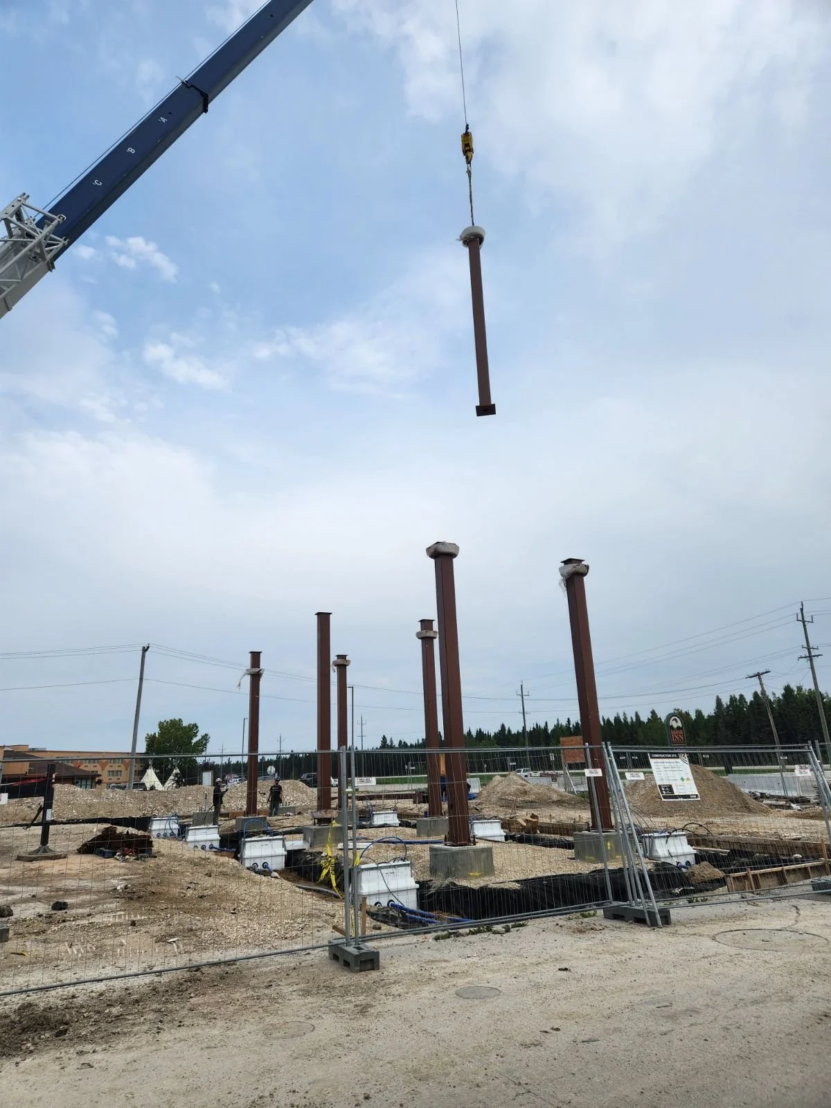 Construction site with multiple steel columns being installed, a crane lifting one column into place, and a fence surrounding the area.