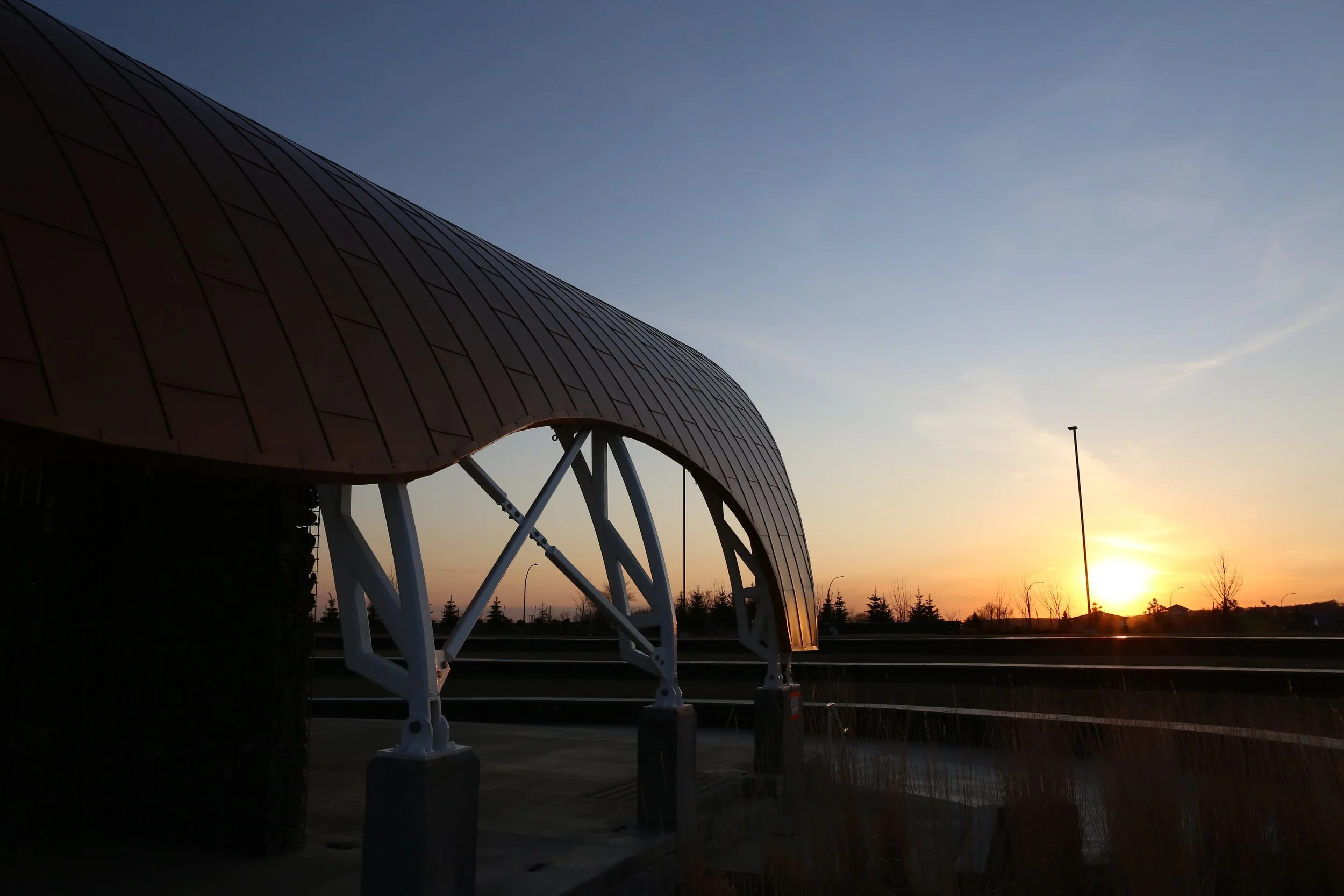 A modern building with curved architecture and metal panels, located beside a highway during sunset, with trees and a lamppost in the background.