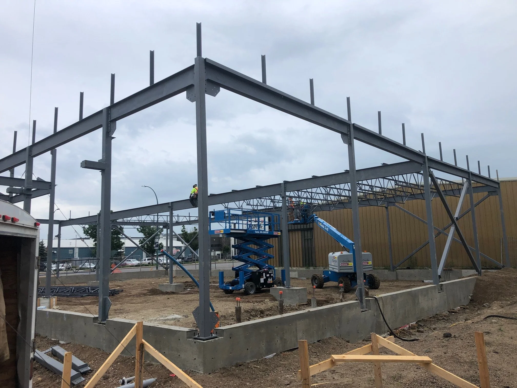 Construction site with steel framework being assembled, with workers and equipment like scissor lifts, on a cloudy day.