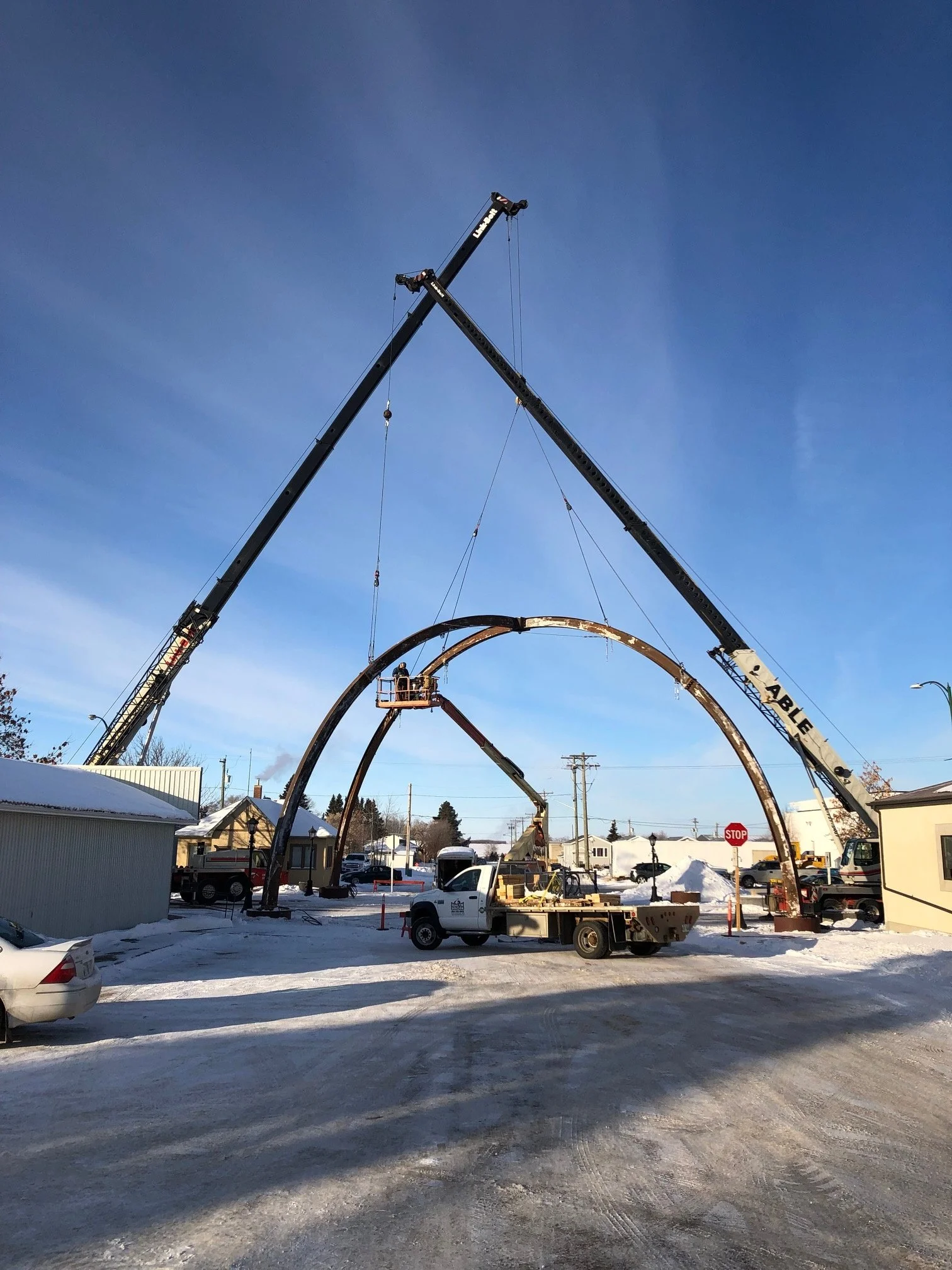 Construction workers are installing a large metal arch structure in a snowy parking lot using cranes and a cherry picker. The scene features a clear blue sky and surrounding buildings.