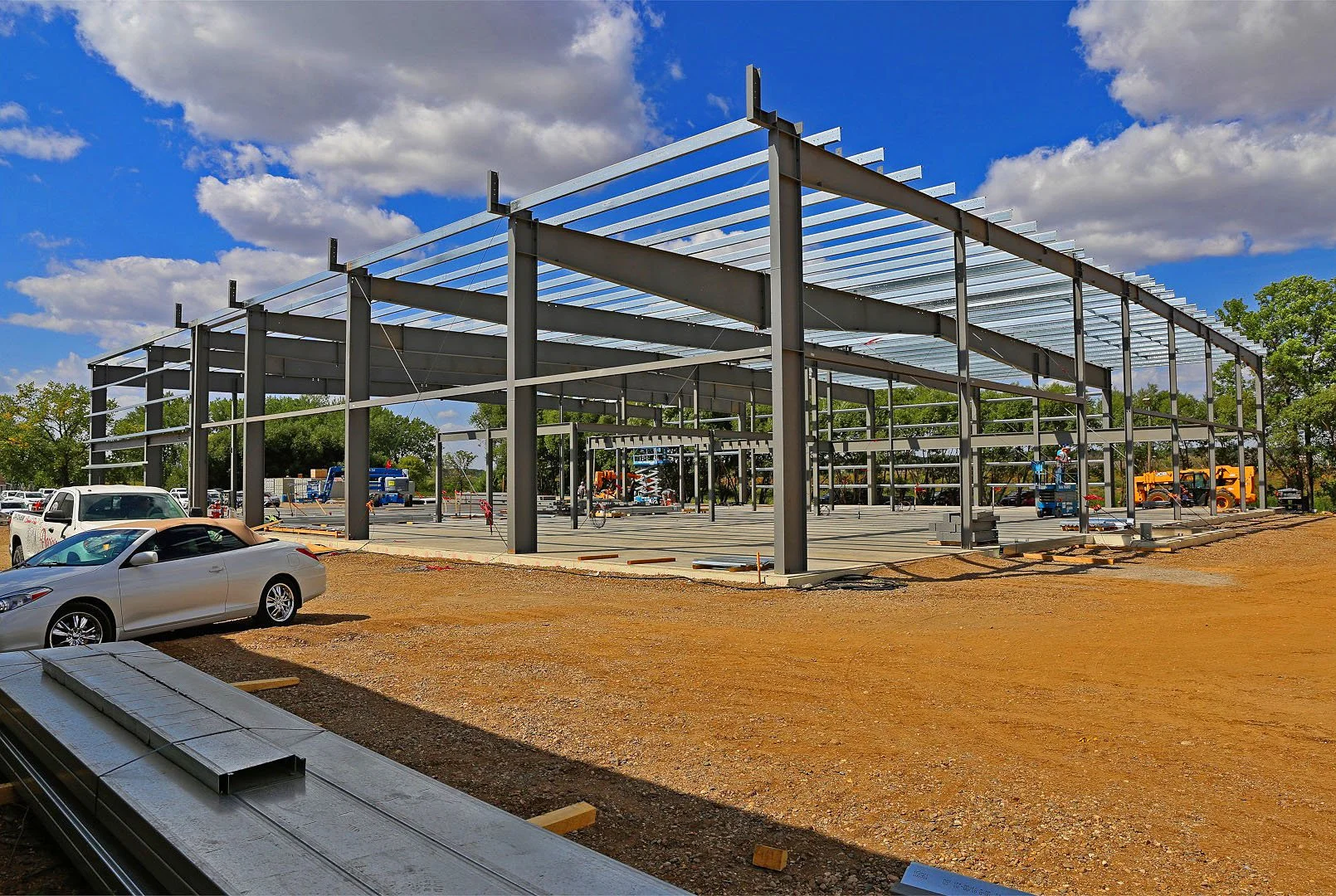 Steel framework of a new building under construction at a construction site on a sunny day with a blue sky and scattered clouds.