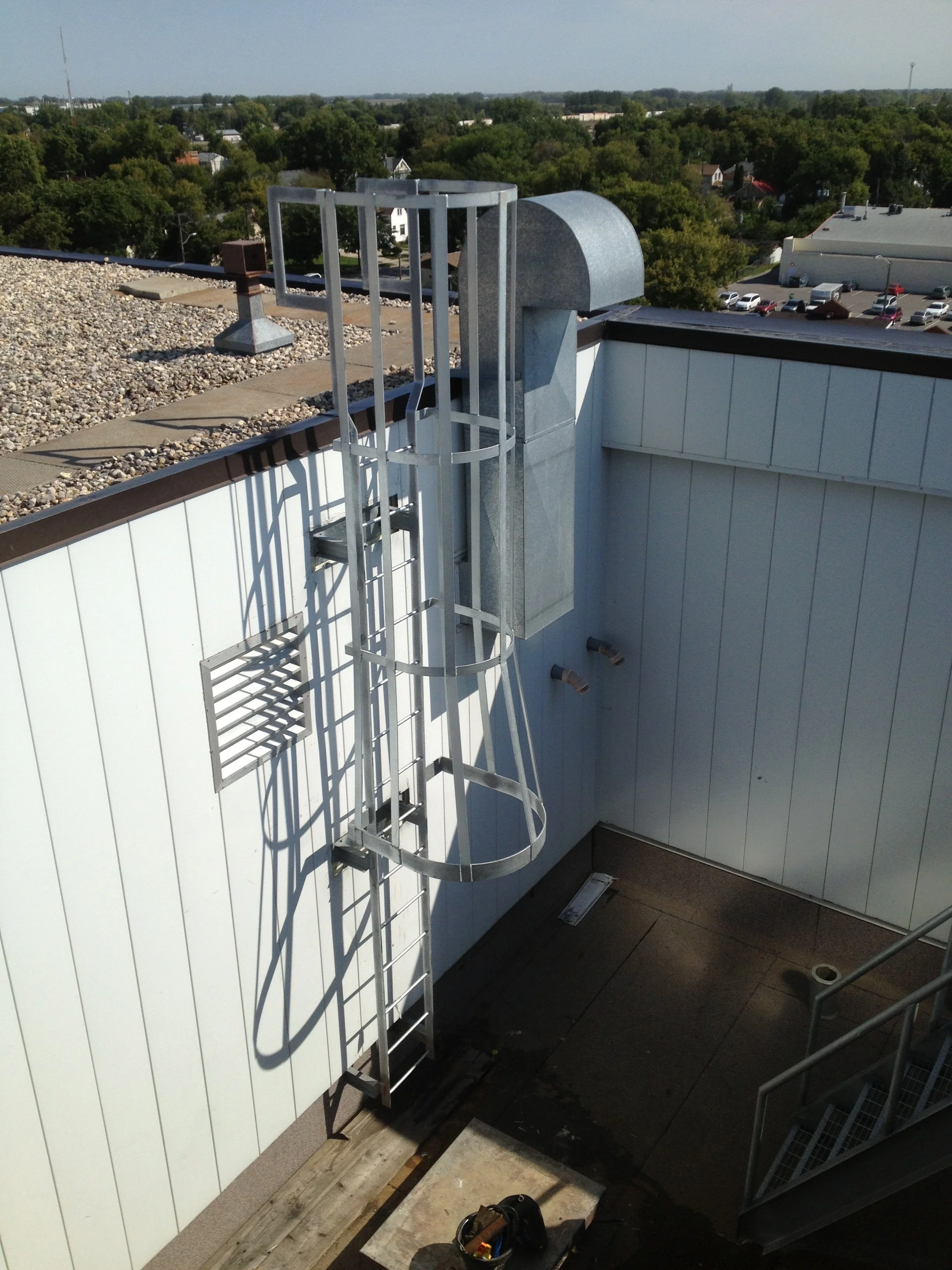 Metal fire escape ladder and vent on the rooftop of a building, with a cityscape and trees in the background.