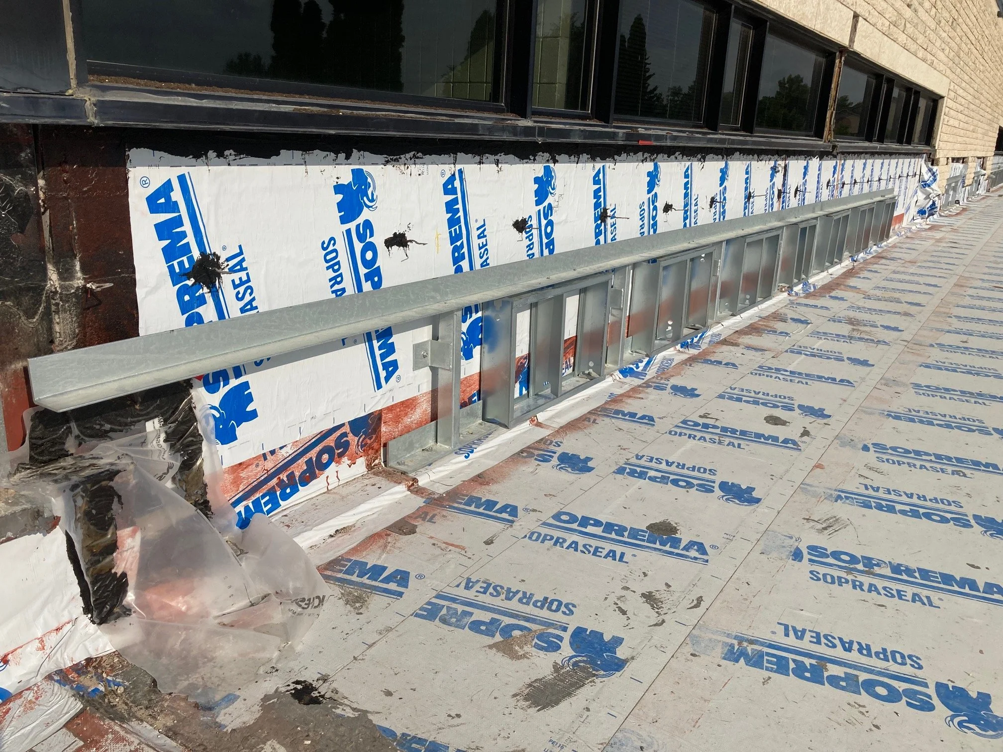 Building exterior under construction with waterproofing and metal framing, waterproofing labeled SOPREMA, on a sidewalk with construction materials.