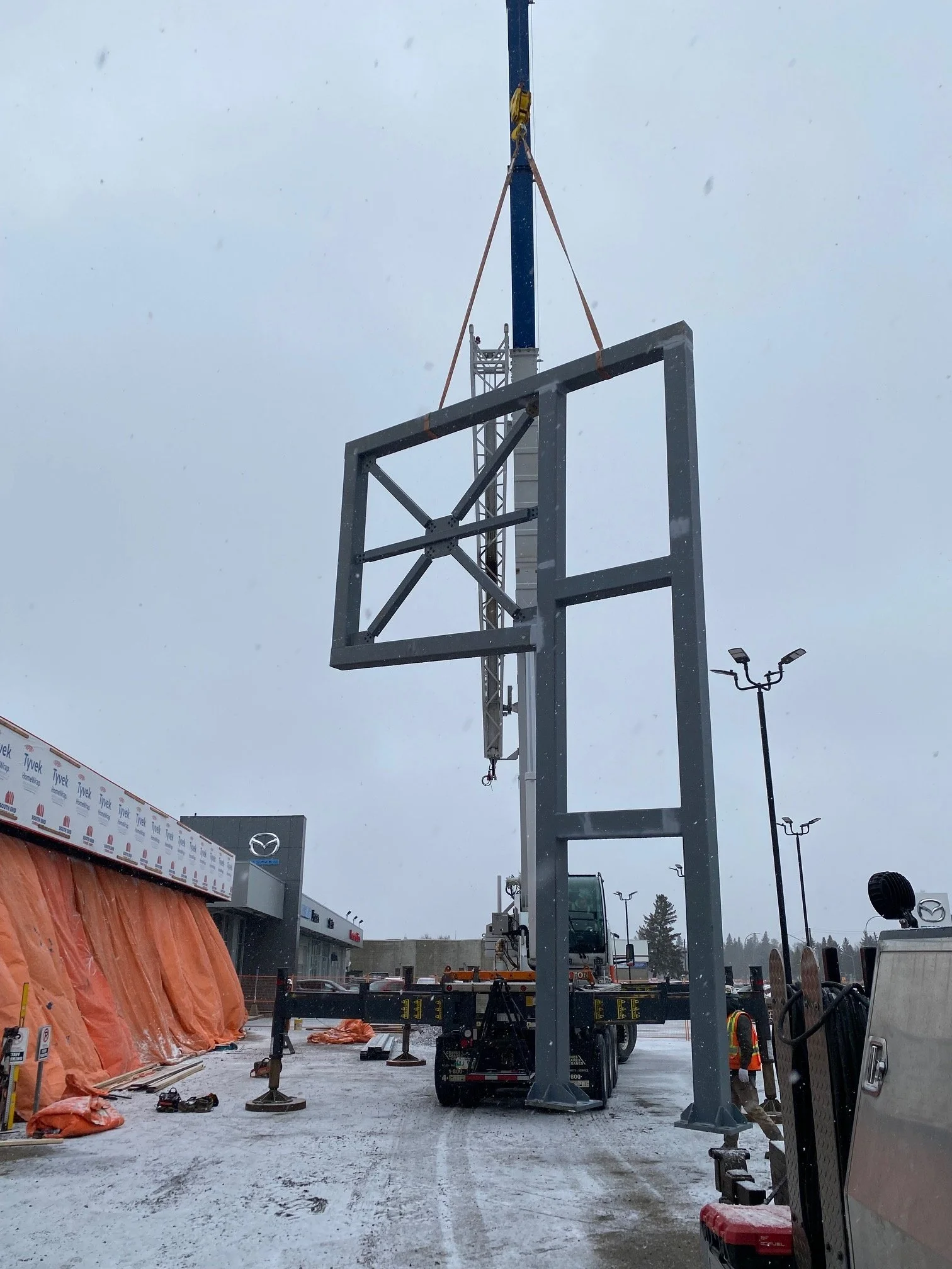Construction site with a crane lifting a large metal frame, snowy ground, orange tarps on the left, and construction workers in safety vests.