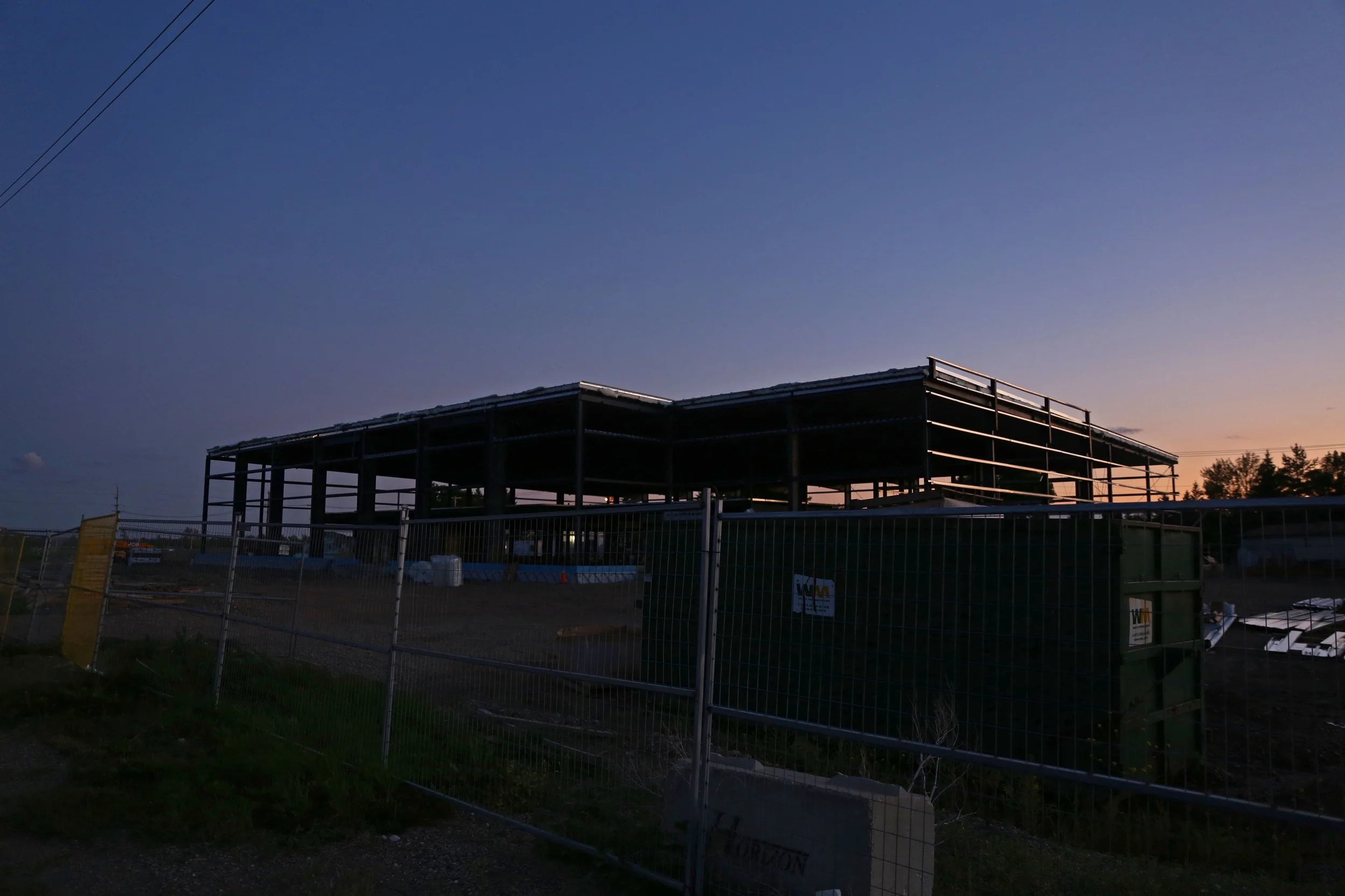 Construction site with a partially built structure at sunset, surrounded by fencing and construction equipment.