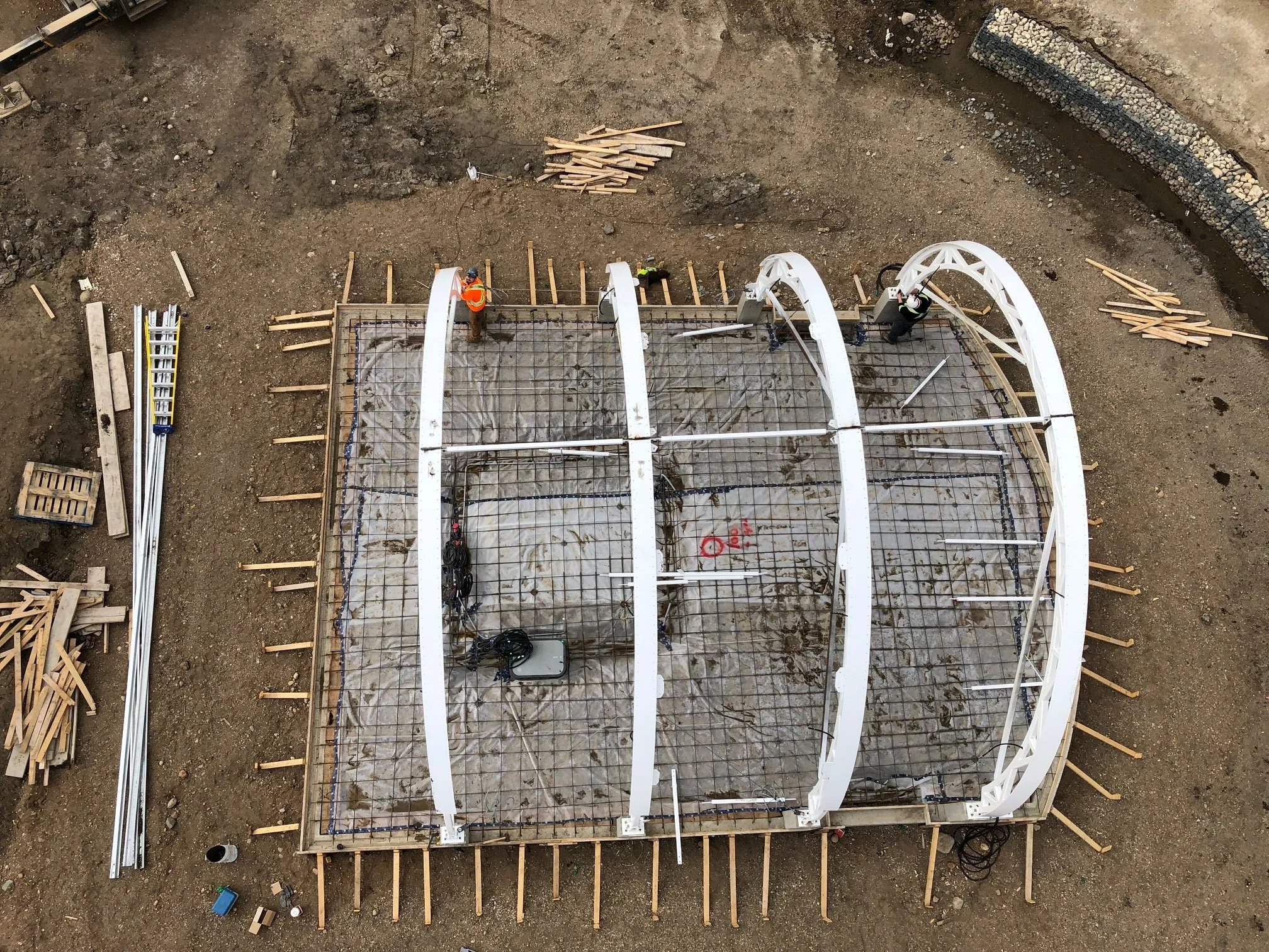 Construction site with a framework of a curved building structure being assembled, workers in safety gear, and scattered construction materials and tools on the ground.