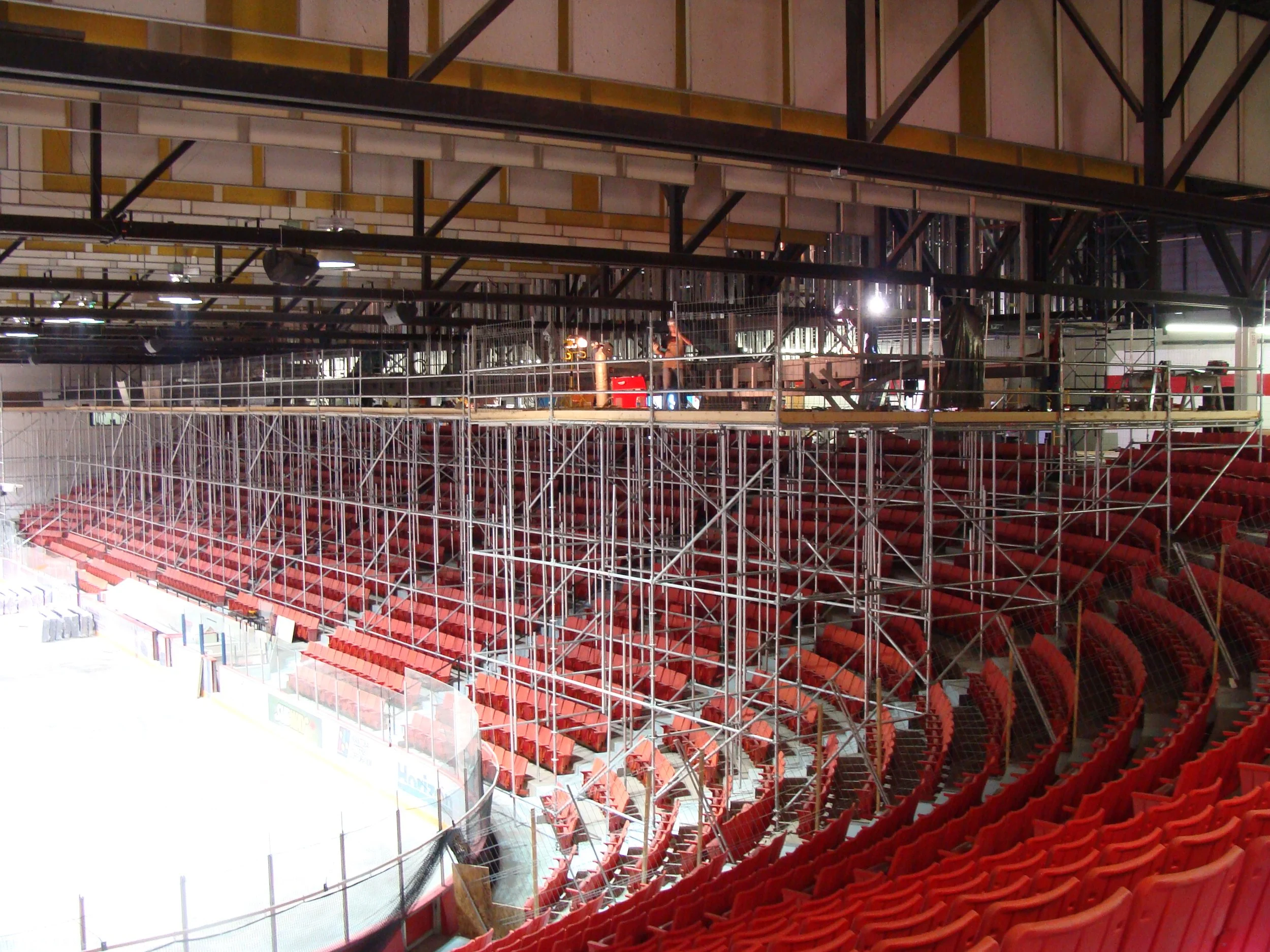 Empty sports arena with red seating and scaffolding construction around the ice rink, indicating a rink upgrade or maintenance.
