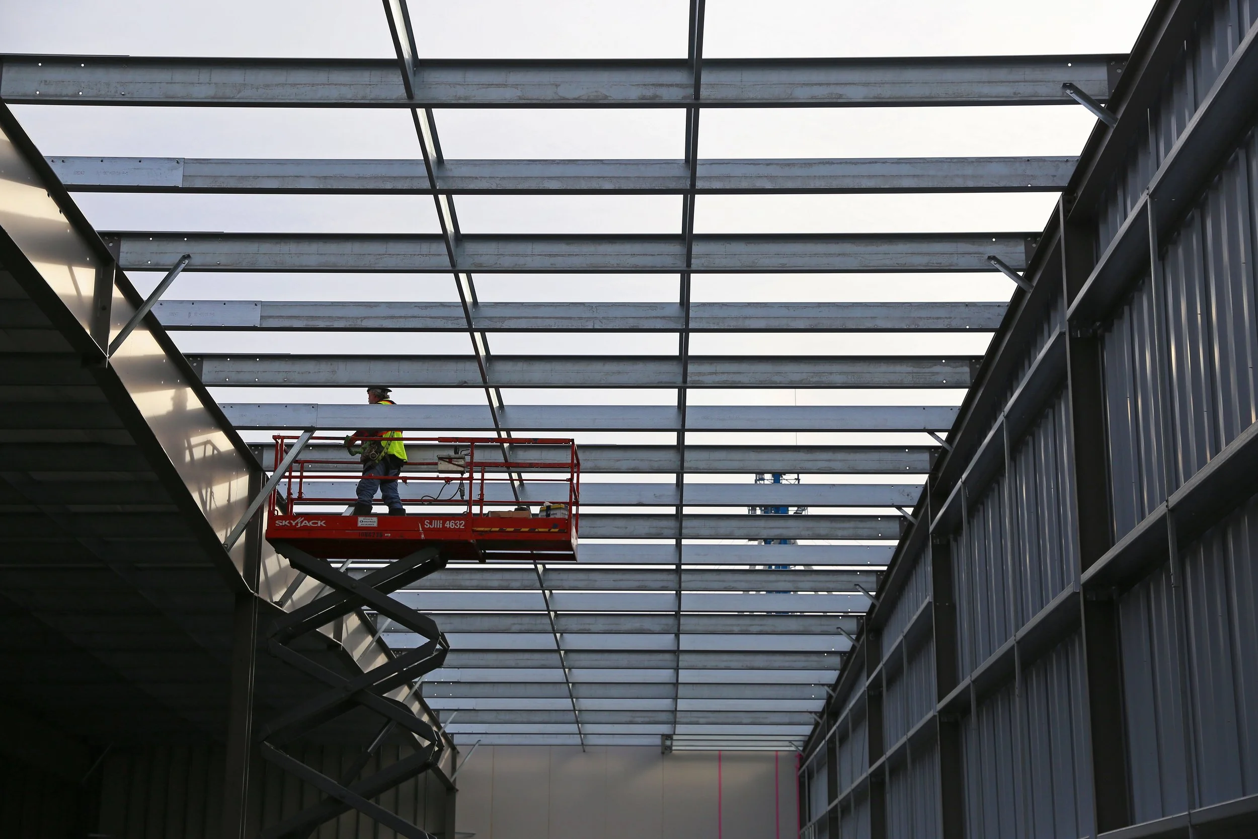 A construction worker in a safety vest and helmet working on a metal framework of a building roof using a cherry picker lift.