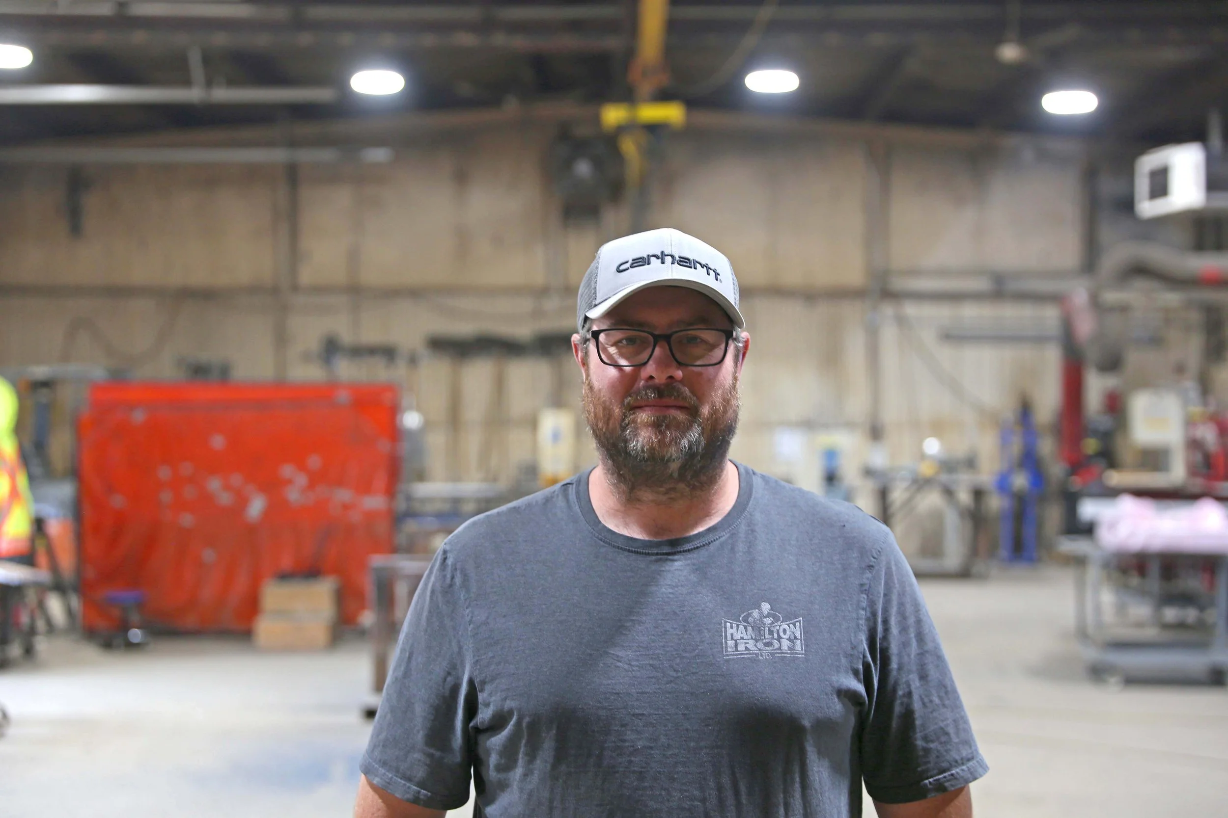 A man with glasses and a beard wearing a gray t-shirt and a white baseball cap with the brand Carhartt logo stands in an industrial workshop or warehouse with equipment and machinery in the background.