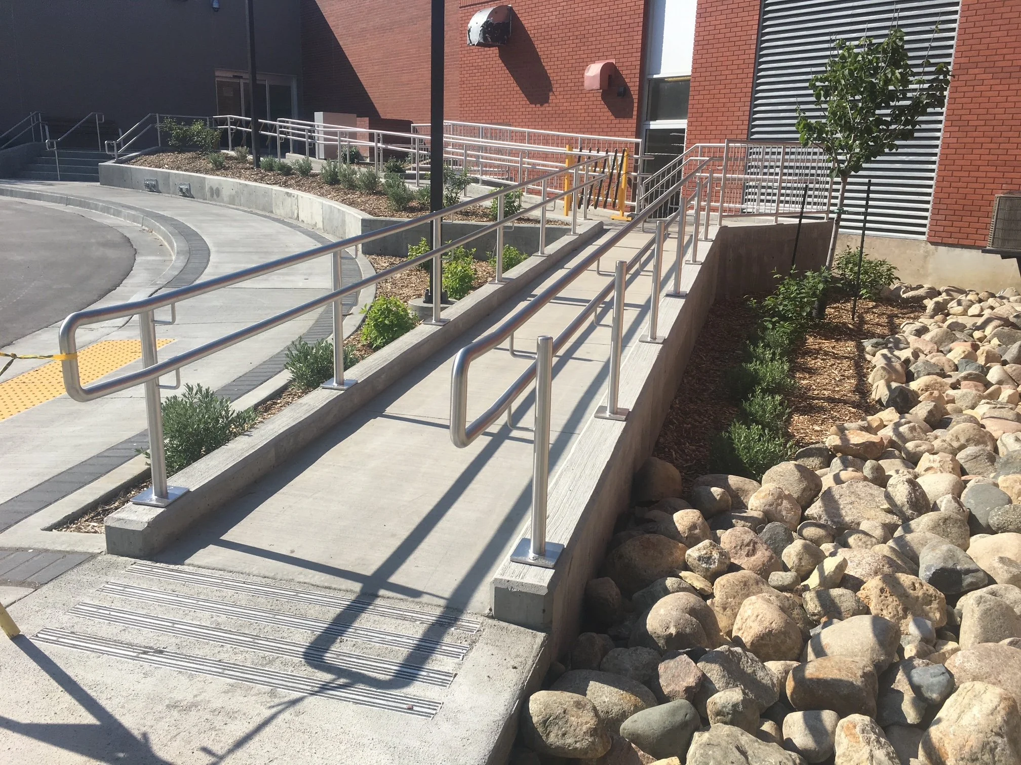 Concrete walkway with handrails on a sloped surface next to a landscaped area with rocks and small plants, adjacent to a brick building.