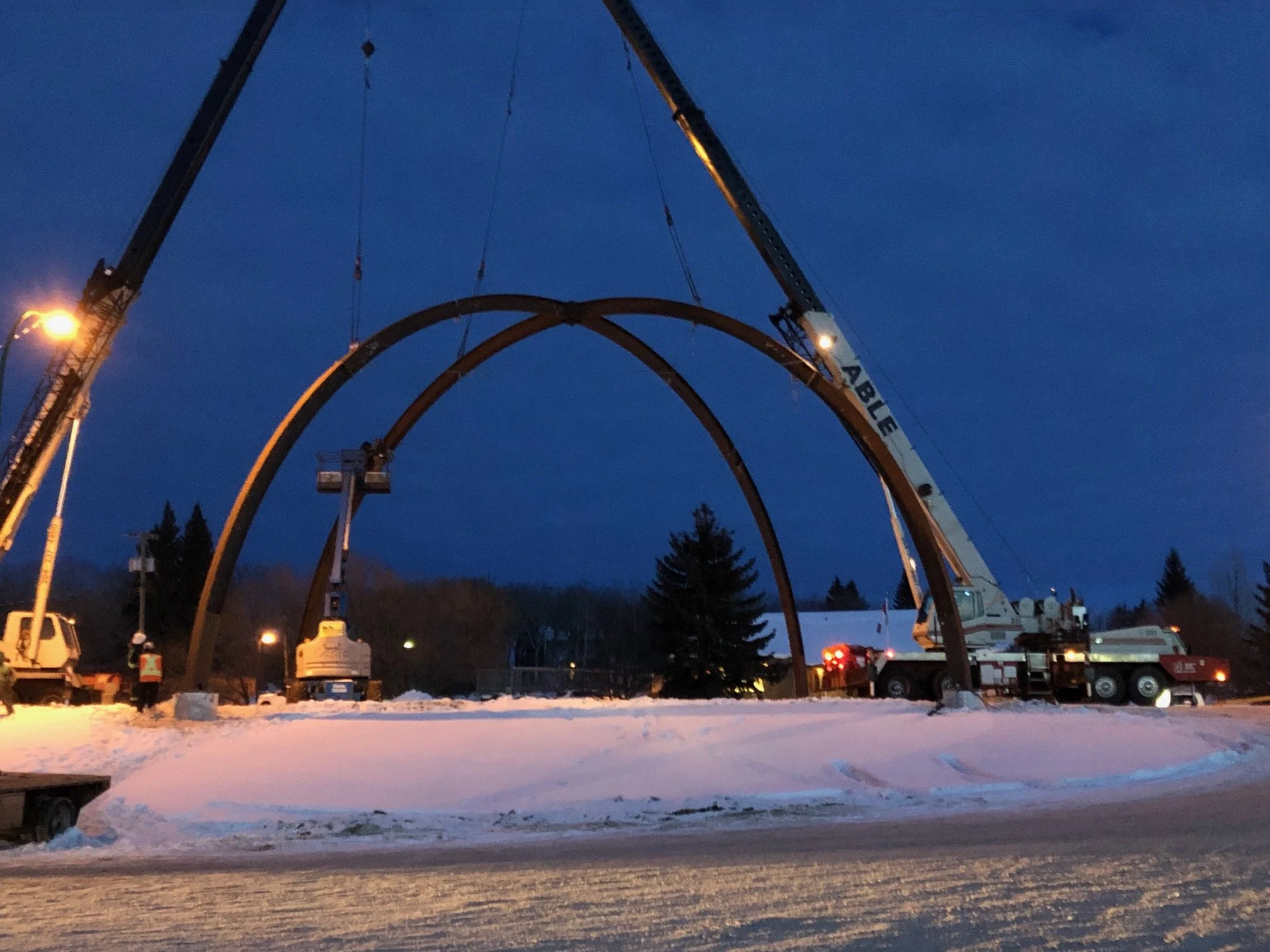 Construction workers using cranes to install large arch structures on a snowy ground during dusk or dawn.