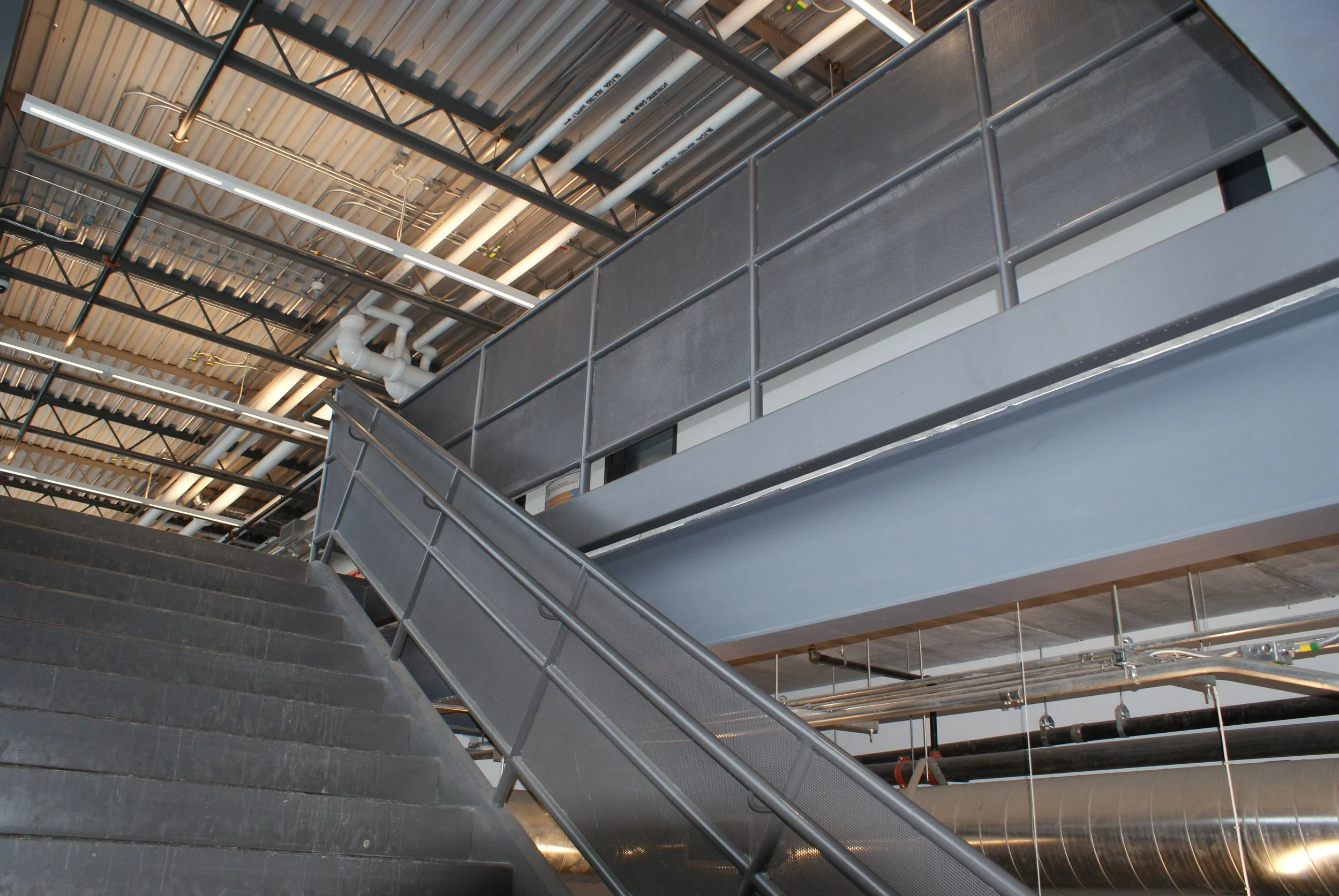 Interior view of an industrial building showing metal stairs, railings, beams, pipes, and ductwork.